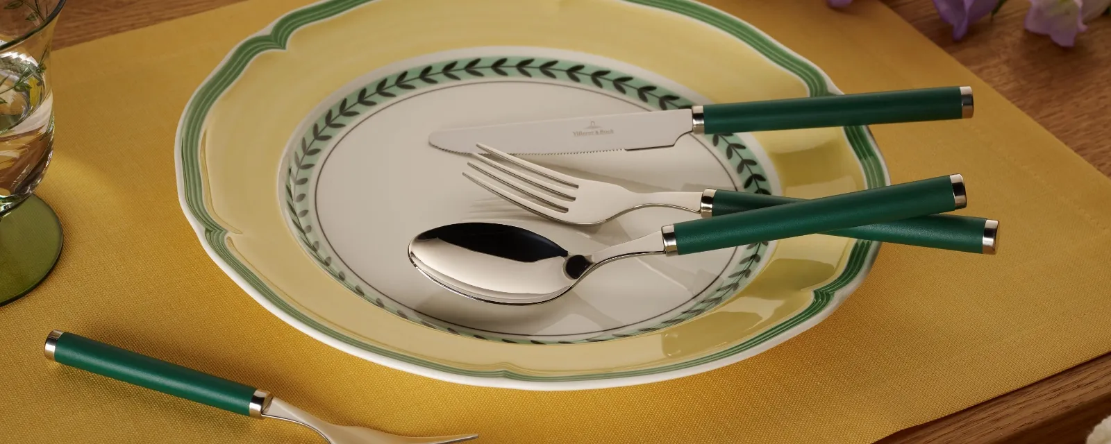 A dining room set is furnished with the Play! collection from Villeroy & Boch and features a decorative plate, flatware with a green handle, and a yellow placemat on a wooden table with purple flowers in the background.