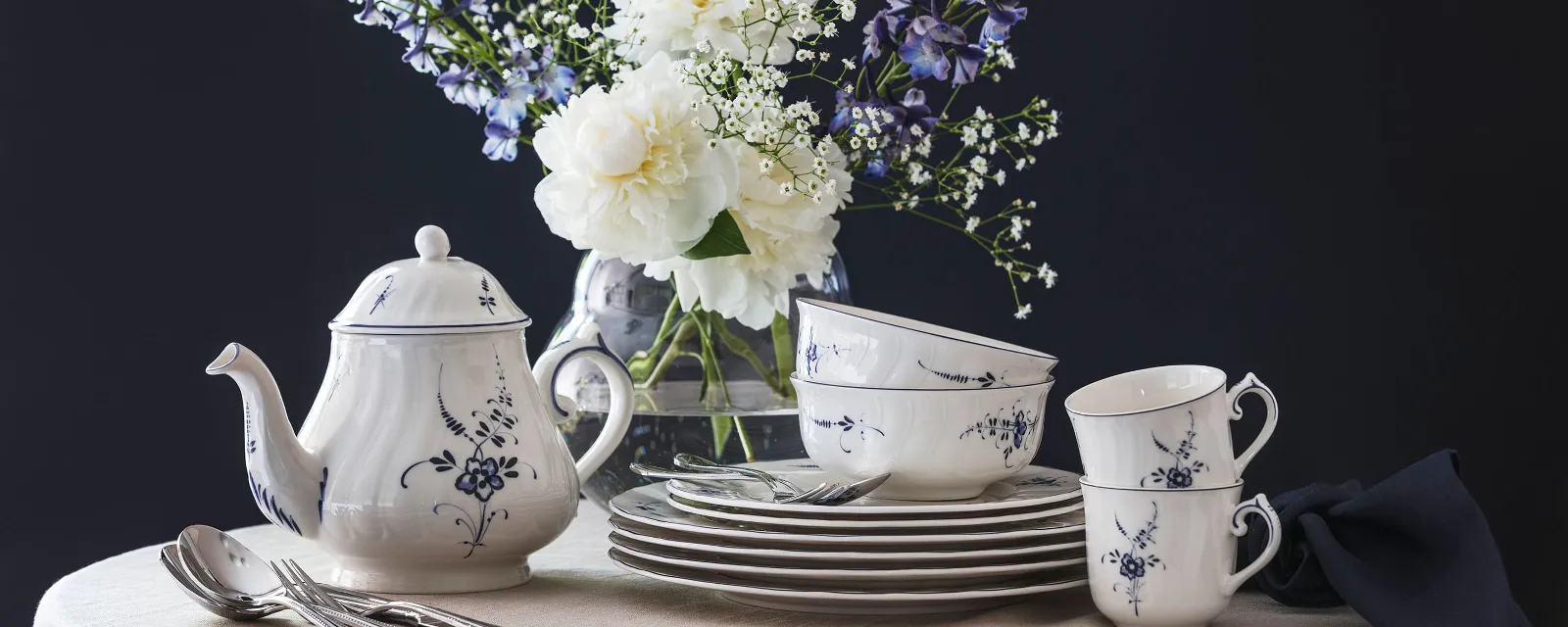 An Old Luxembourg porcelain set with a blue and white floral pattern, consisting of a teapot, cups and plates on a round table, with a vase of white and purple flowers in the background.