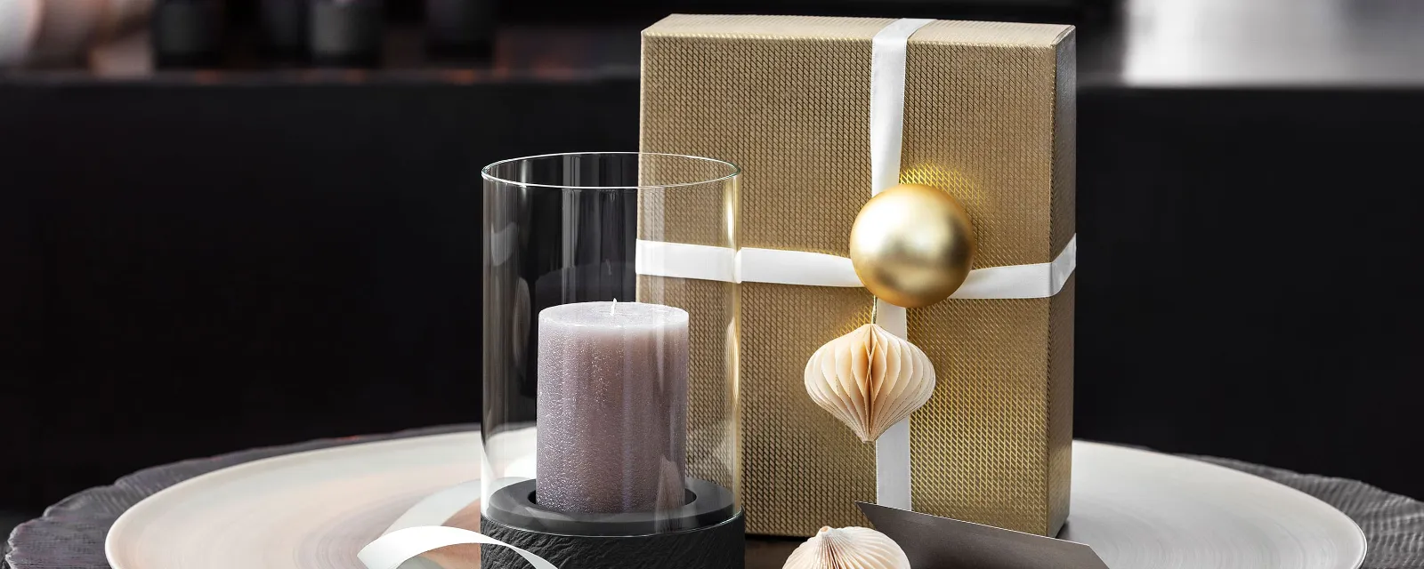 A Manufacture hurricane lamp with a candle next to a gift wrapped with a white box and ornaments.