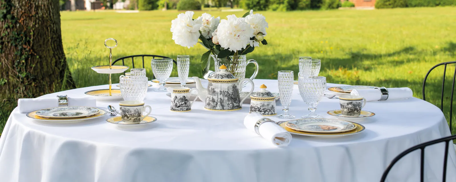 Elegant outdoor dining with fine Audun place settings and Boston glasses on a white tablecloth in front of a historic castle on a sunny day.