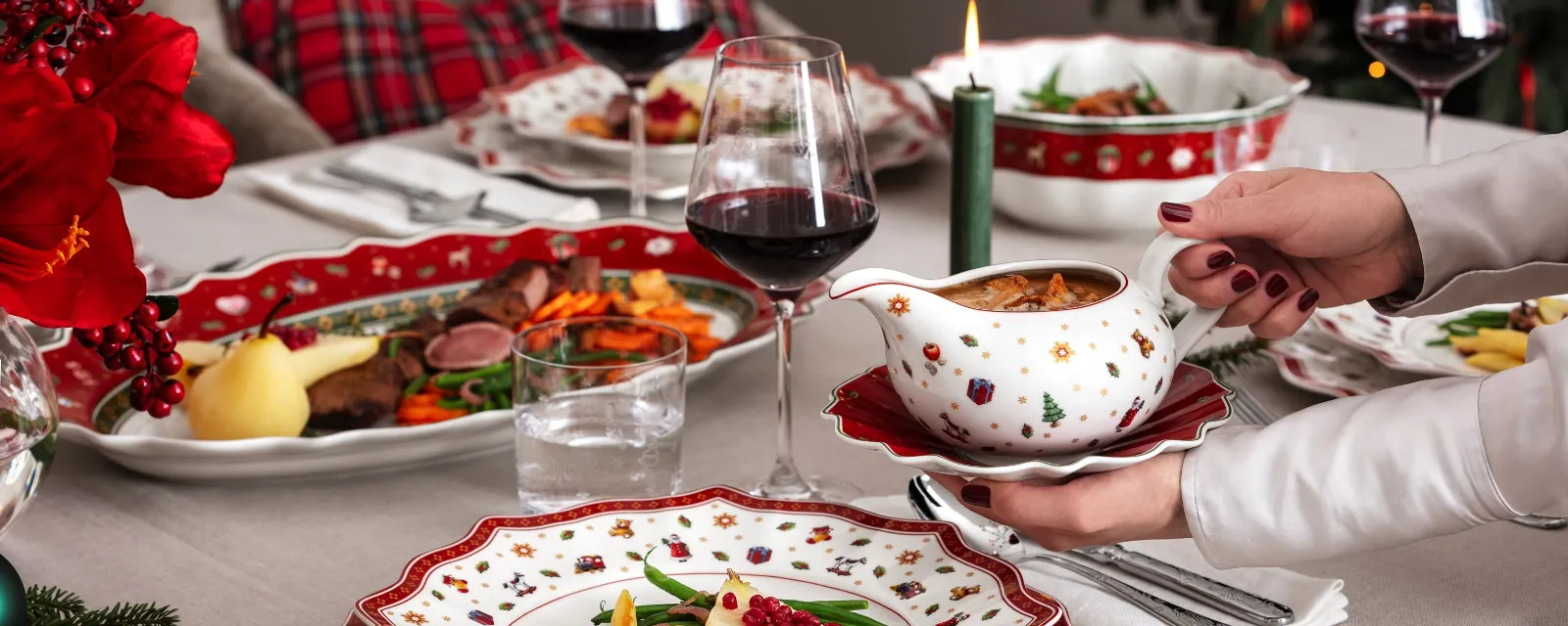 A festive dining table setting with Christmas dinnerware from Villeroy & Boch Toy’s Delight and presents plates of meat, vegetables, and sauce. A person holds a gravy boat, glasses of red wine, and a lighted candle.