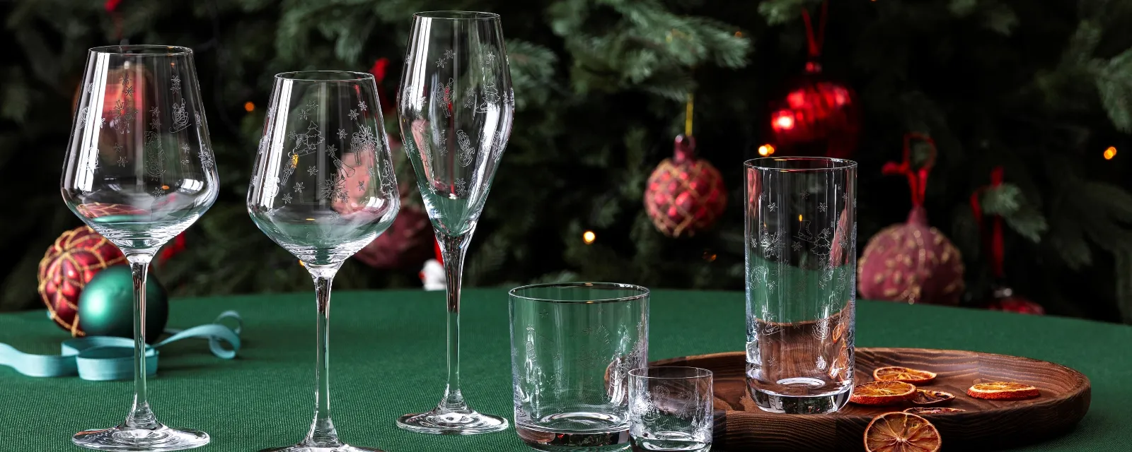 A stylish arrangement from the Toys Delight glassware collection by Villeroy & Boch, consisting of wine glasses and drinking glasses, elegantly placed on a green table. Dried citrus slices lie on a wooden tray in the foreground, complemented by the festive presence of a decorated Christmas tree in the background.