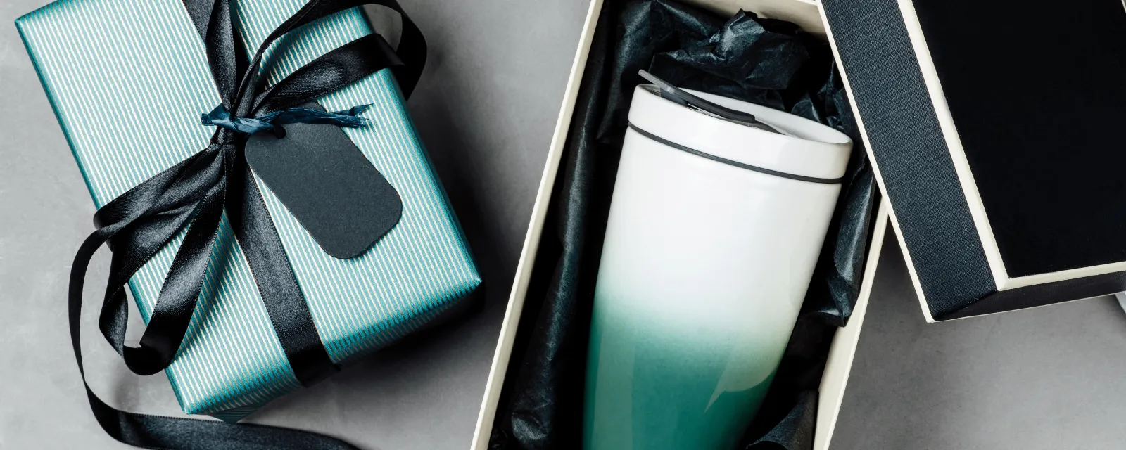 A ceramic “To go and To stay” travel mug from Villeroy & Boch with a color gradient from white to blue-green is presented in an open gift box lined with black tissue paper, while next to it is a closed gift box wrapped in a black bow.