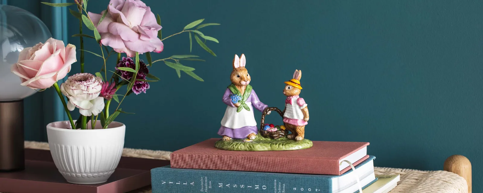 Two decorative bunny figurines from the Villeroy & Boch Bunny Tales collection on stacked books on a woven bench, next to a vase with buttercups and roses.