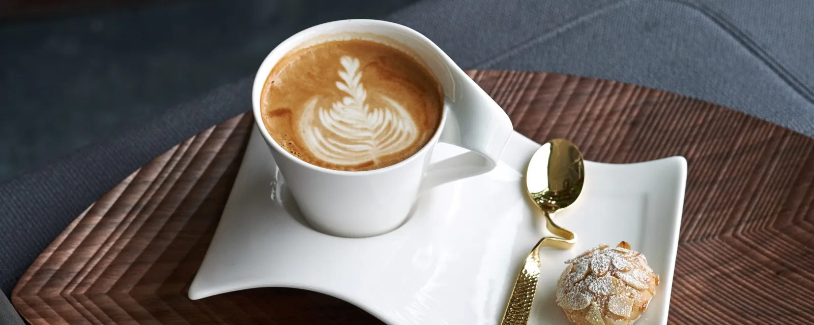 A Villeroy & Boch NewWave latte cup with latte art on a wavy white saucer is combined with a small cookie and a golden spoon, all beautifully arranged on a wooden tray.