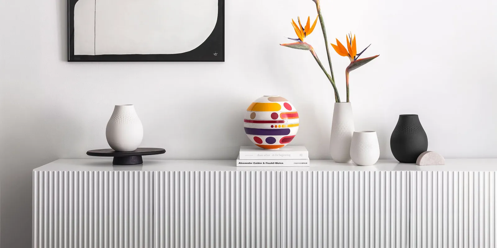 A white sideboard with vases, the colorful La Boule Miami and books underneath, next to a bird of paradise flower arrangement, with geometric wall art in the background.