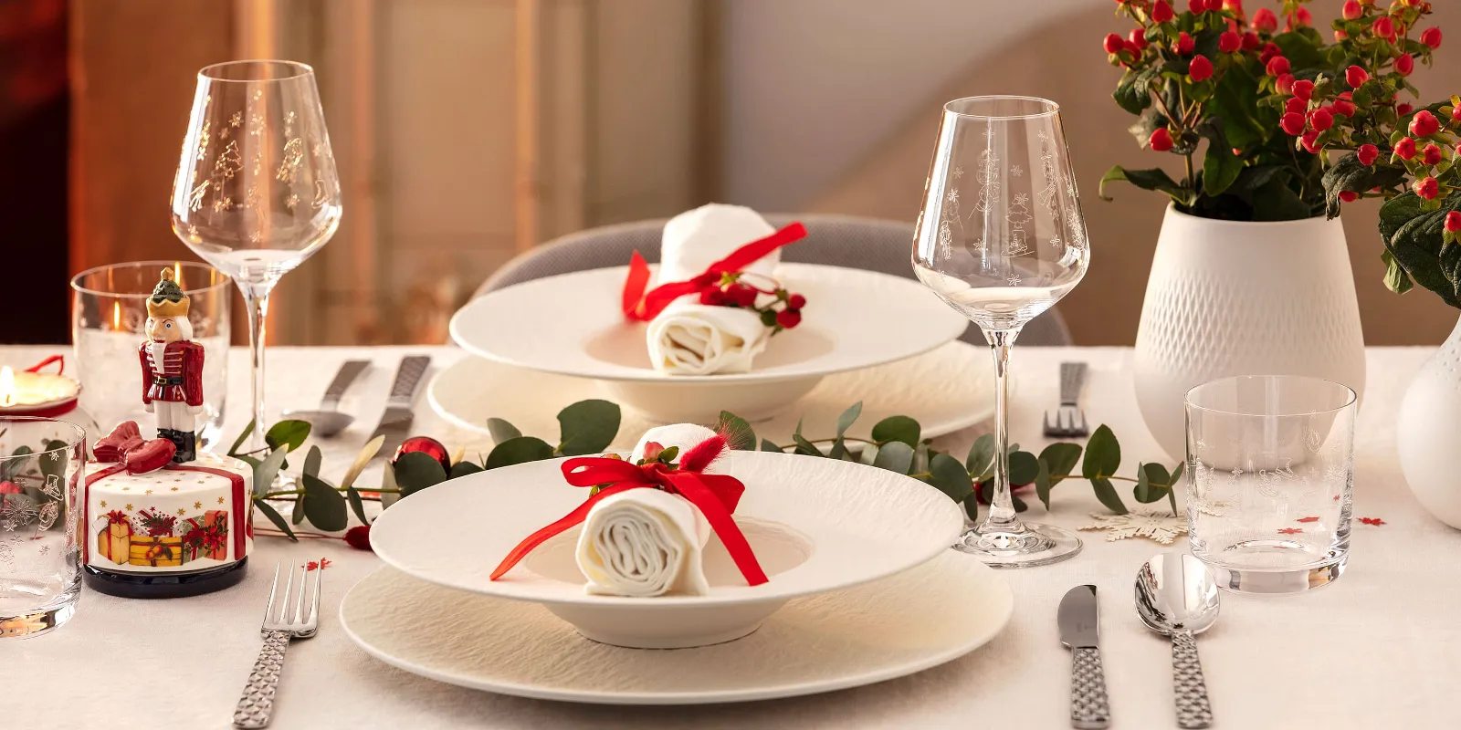 Elegant dining table for two with white Manufacture plates, napkins with red ribbons, wine glasses, silver flatware, and decorative plants.