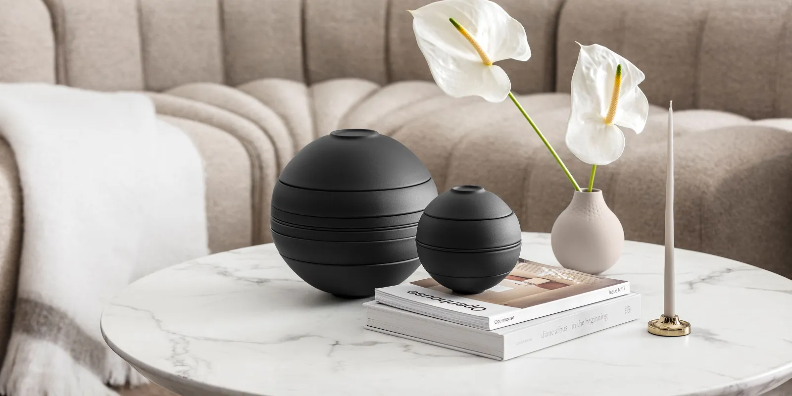 The Villeroy & Boch black La Boule, a beige vase with white flowers, a candleholder and some books in a beige living room setting.