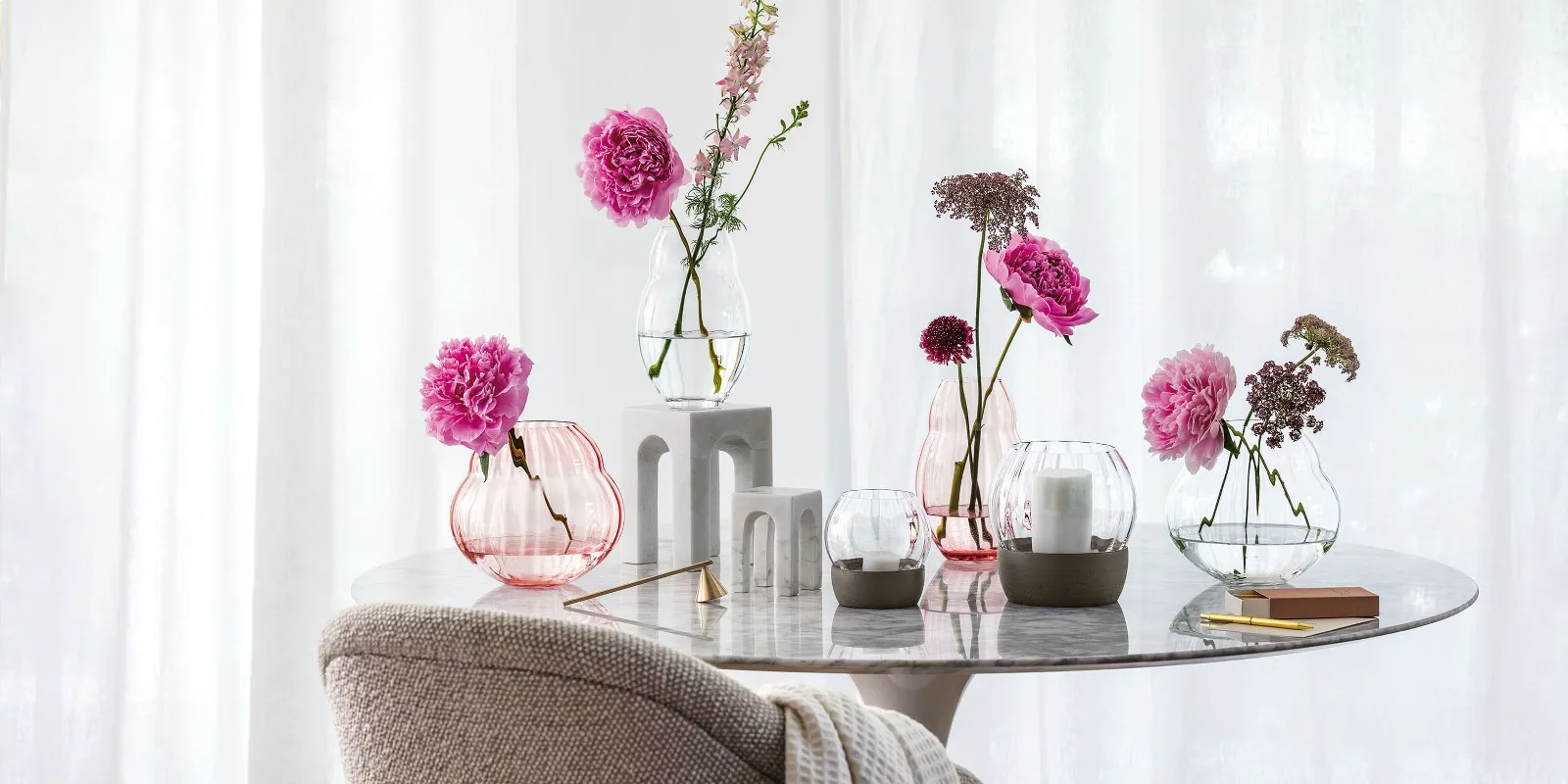 A marble table with various flowers in Rose Garden glass vases and Rose Garden hurricane lamps in front of a window with curtains.