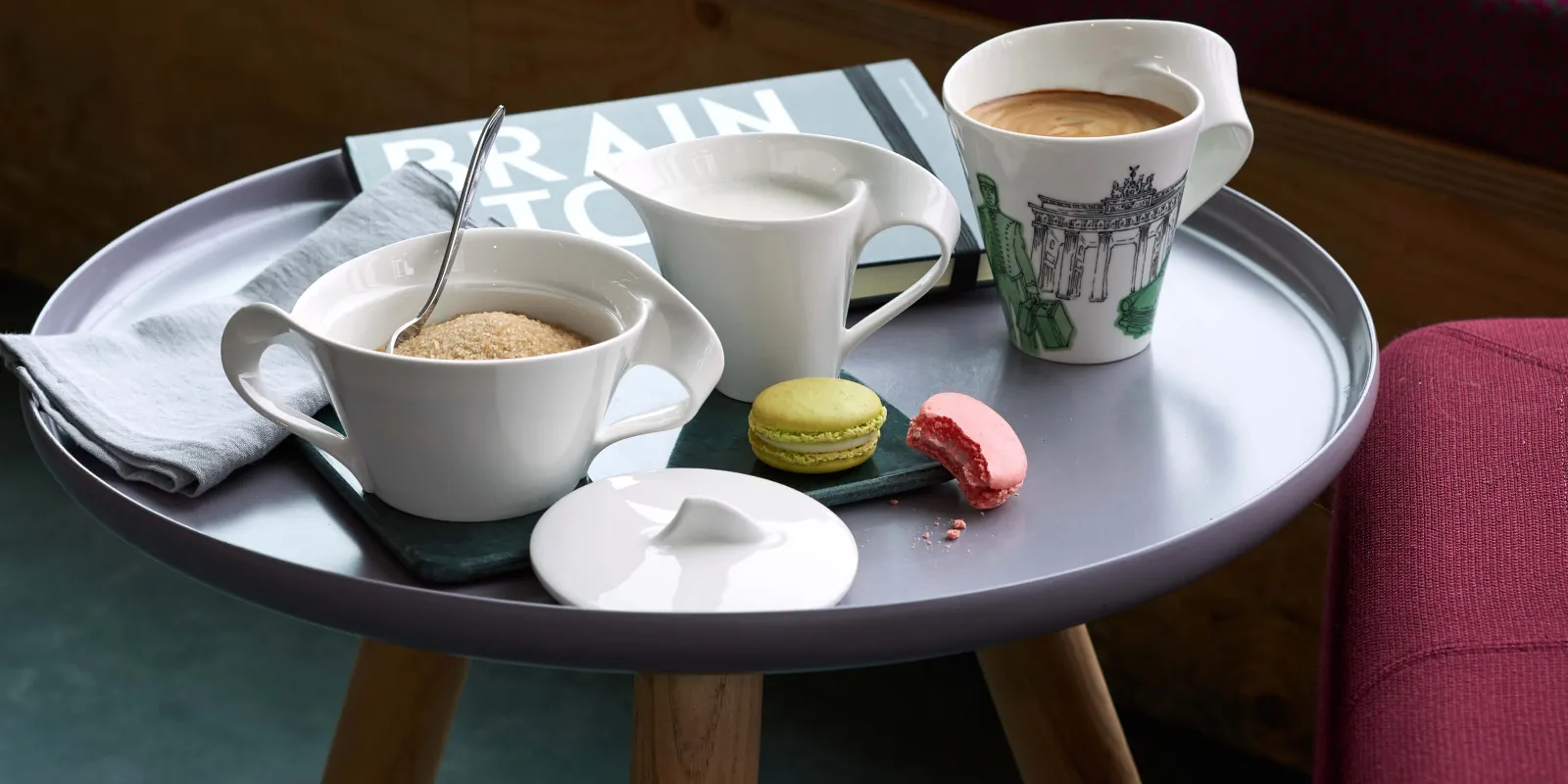 NewWave coffee cup, a milk jug, a sugar bowl with a spoon and two macarons on a saucer, on a small table.