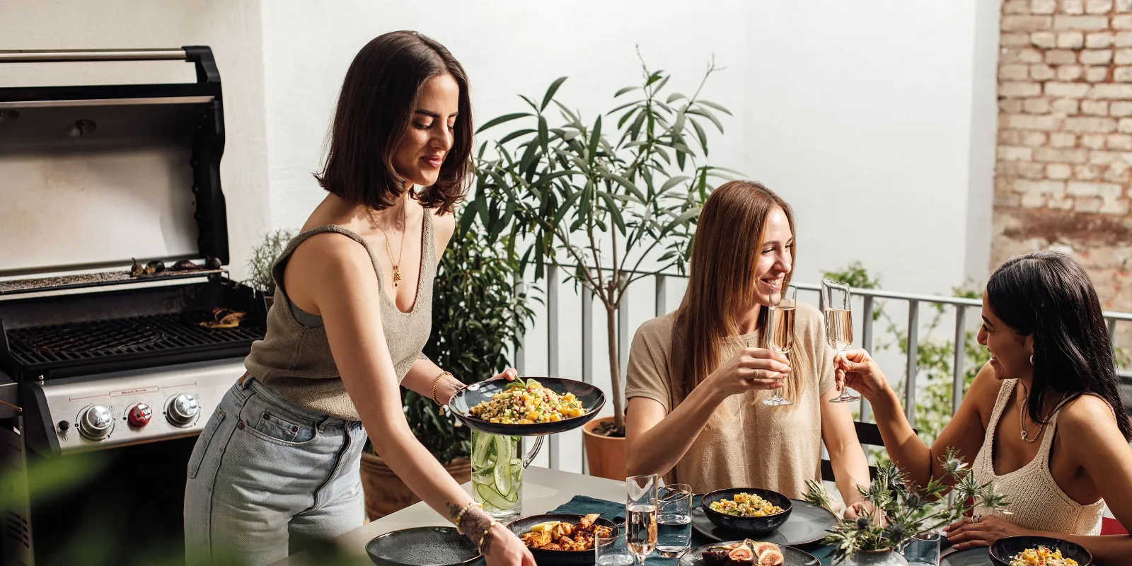 Two friends enjoy summer delicacies on Lave plates and toast with a glass of sparkling wine, another person serves food.