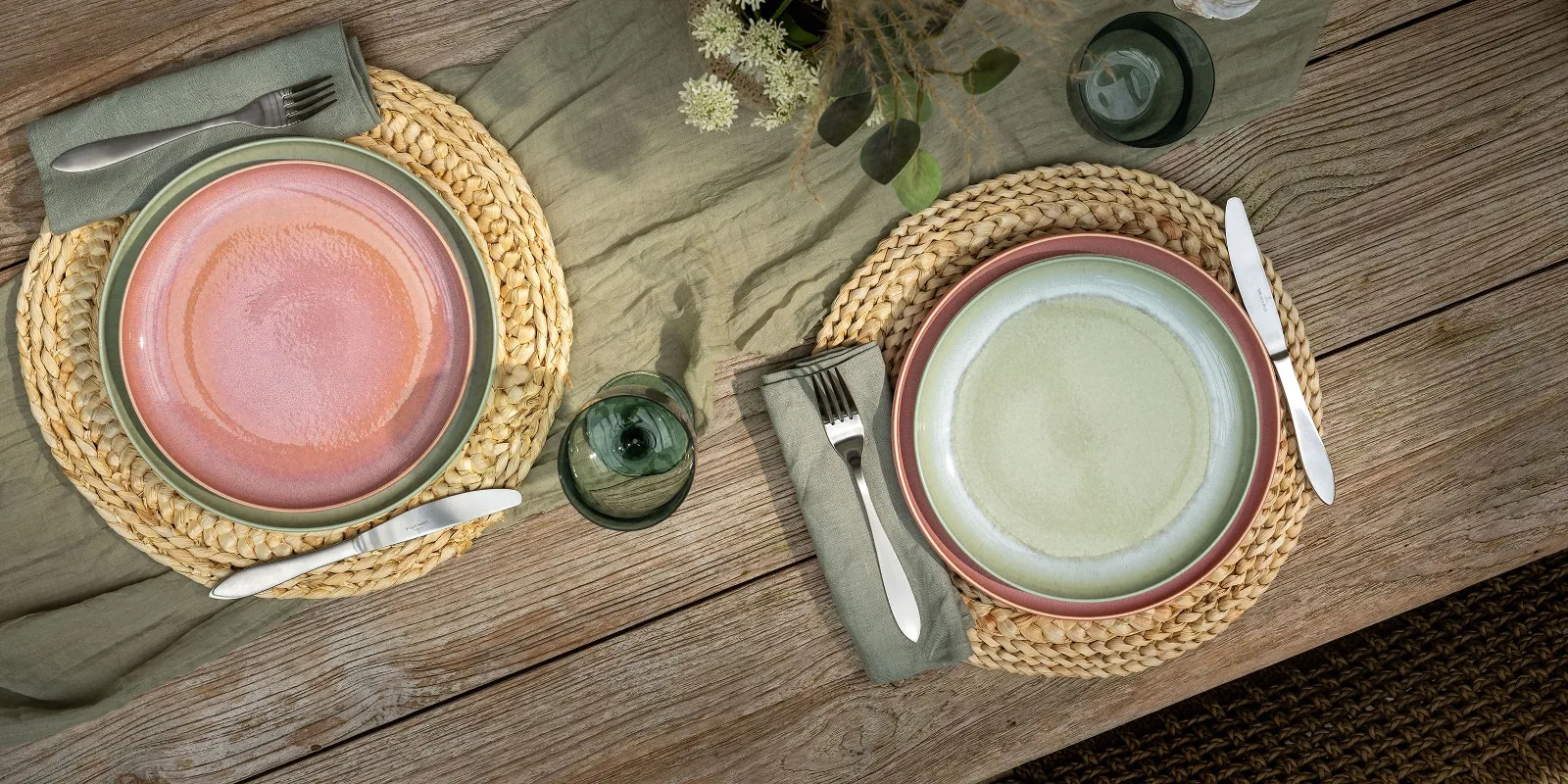 Two Perlemor place settings on a wooden table with round woven placemats, cutlery, a candle, flowers and green plants.