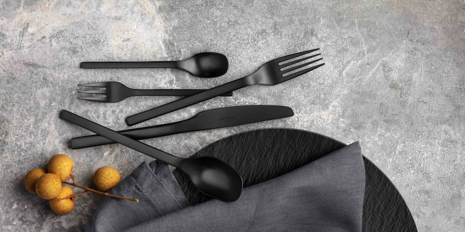 Black Manufacture cutlery set with a fabric napkin and decorative berries on a textured grey surface.