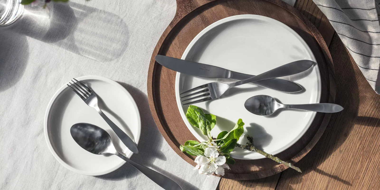 Elegant table decoration with simple white plates and Arthur Brushed stainless steel cutlery, accentuated by natural light and a sprig of flowers.