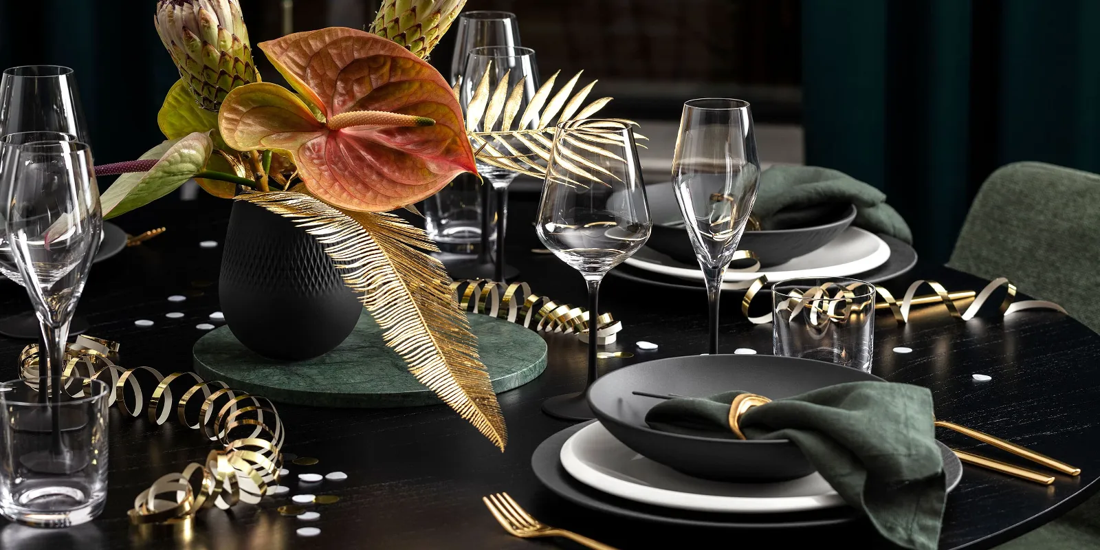 Elegant Villeroy & Boch table setting with dark Manufacture plates, gold cutlery, green napkins and a black Collier vase with tropical flowers and leaves. Gold streamers and glasses decorate the black table.