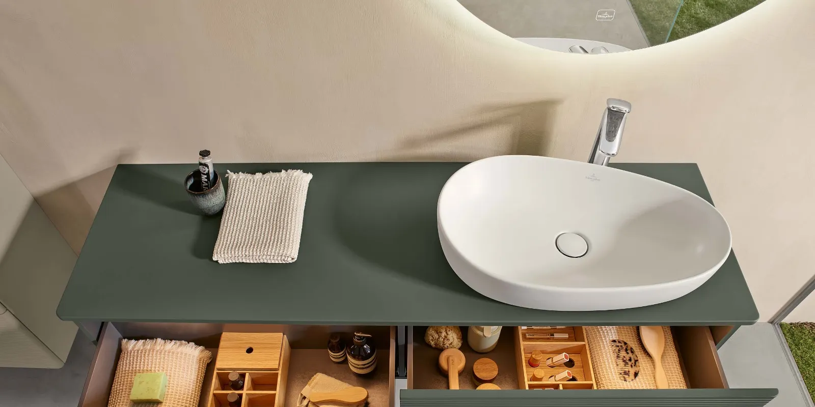 A natural green console in a bathroom with two open, full drawers, above which is a freestanding white washbasin from Villeroy & Boch Antao.