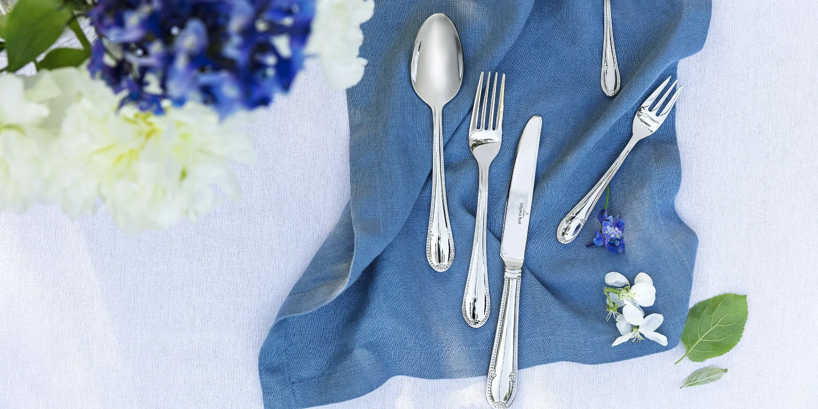 Stylish table decoration with Mademoiselle cutlery on a blue linen napkin and a bouquet of flowers.