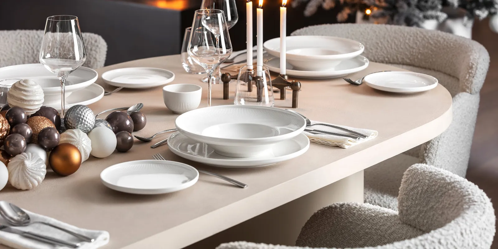 Ein festlicher Esstisch mit weißem Afina-Geschirr, Kerzen und dekorativen Ornamenten von Villeroy &amp; Boch, neben einem brennenden Kamin und einem geschmückten Weihnachtsbaum im Hintergrund.