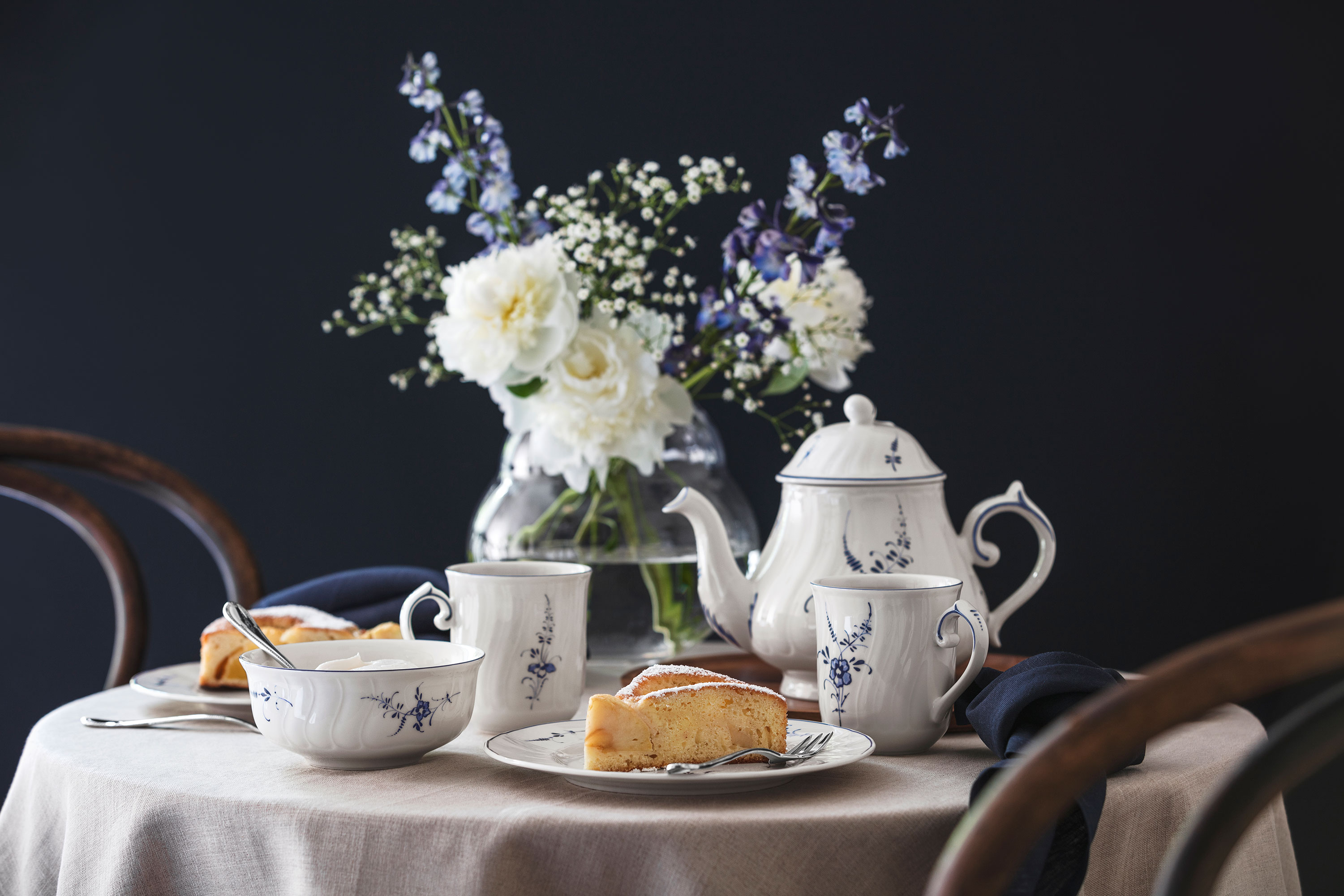 A round table set with vieuy luxembourg tea, teacups, cake slices, and a vase of white flowers against a dark background.
