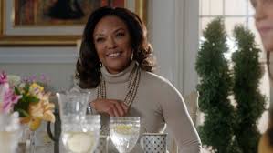 greenleaf-as-seen-on A woman in a beige turtleneck sits at a table set with flowers and water glasses, smiling warmly. The setting is elegant and softly lit, conveying a welcoming atmosphere.
