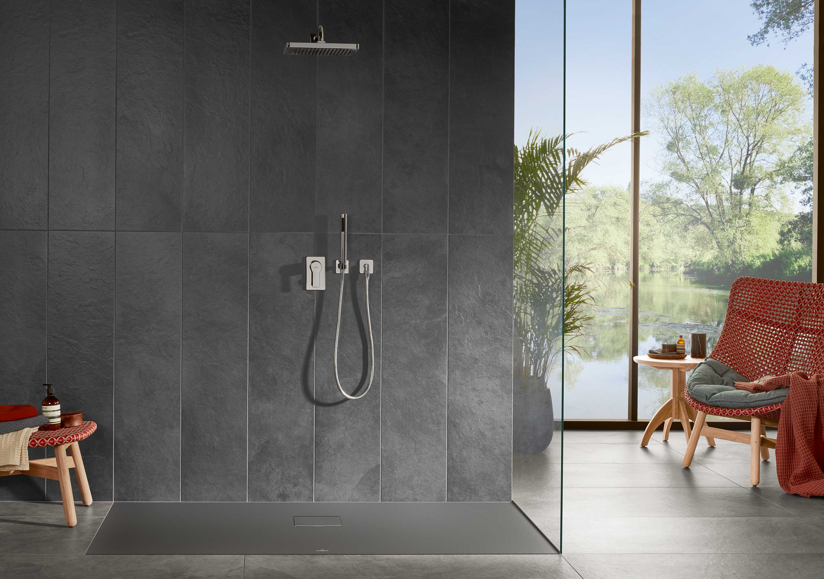 Un espace douche moderne avec le receveur de douche Squaro Infinity de Villeroy & Boch Un espace douche moderne avec du carrelage gris foncé, un receveur de douche Squaro Infinity de Villeroy & Boch, une pomme de douche à effet pluie et douchette à main. Un coin détente adjacent avec chaises en osier rouge et vue sur l’extérieur à travers des parois vitrées.