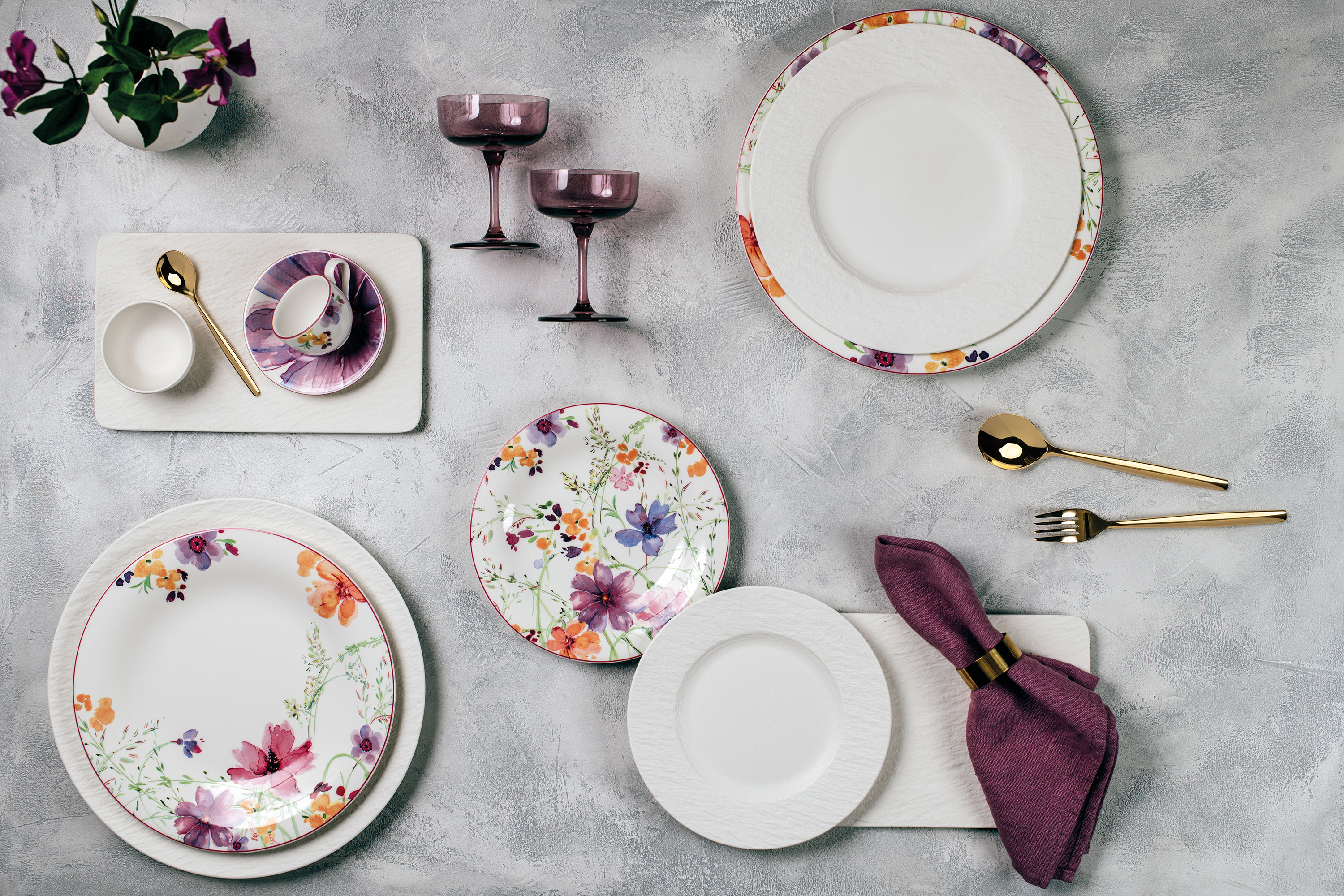 Een verfijnde tafelopstelling bestaat uit borden met bloemmotief "Mariefleur" van Villeroy & Boch. Een verfijnde tafelopstelling bestaat uit borden met bloemmotief "Mariefleur" van Villeroy & Boch, gouden bestek, paarse servetten en bijpassend glaswerk op een gestructureerd grijs oppervlak.