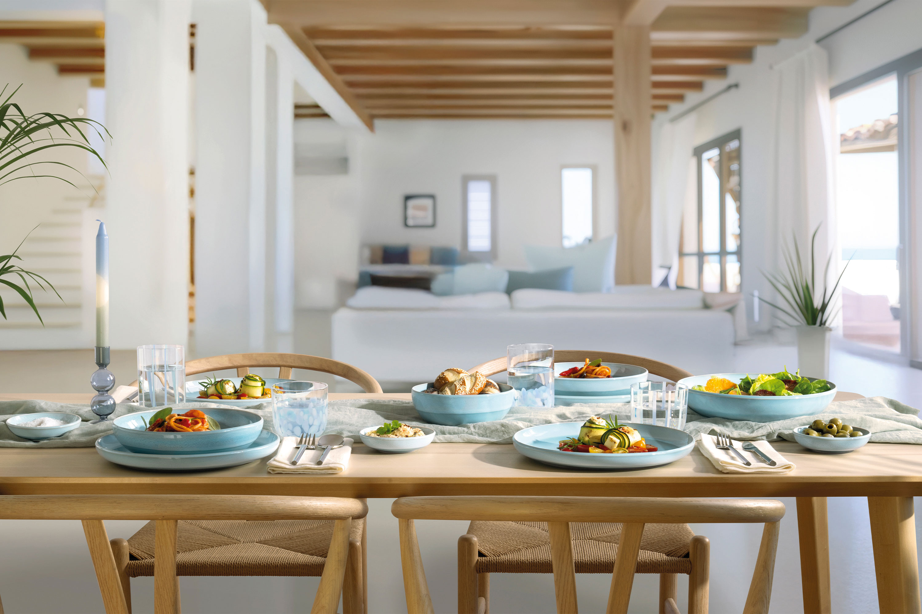 A wooden dining table for four people with blue Perlemor Aqua plates A wooden dining table for four people with blue Perlemor Aqua plates in a bright, modern living area.