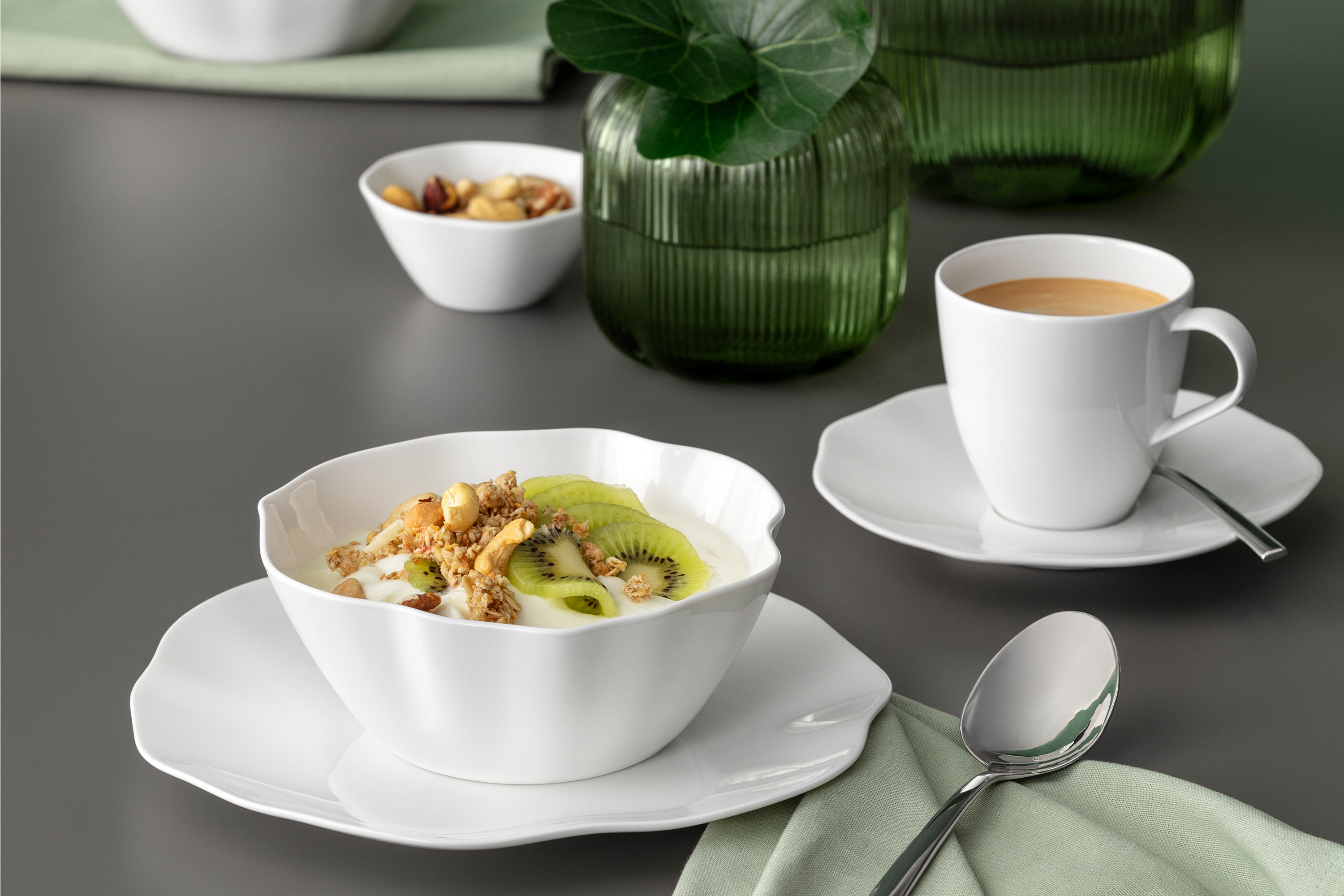 Eine weiße Pura Schale mit Müsli und Kiwi und eine Tasse Kaffee auf einer Untertasse Eine weiße Pura Schale mit Müsli und Kiwi und eine Tasse Kaffee auf einer Untertasse auf einem grauen Tisch mit einer grünen Serviette.