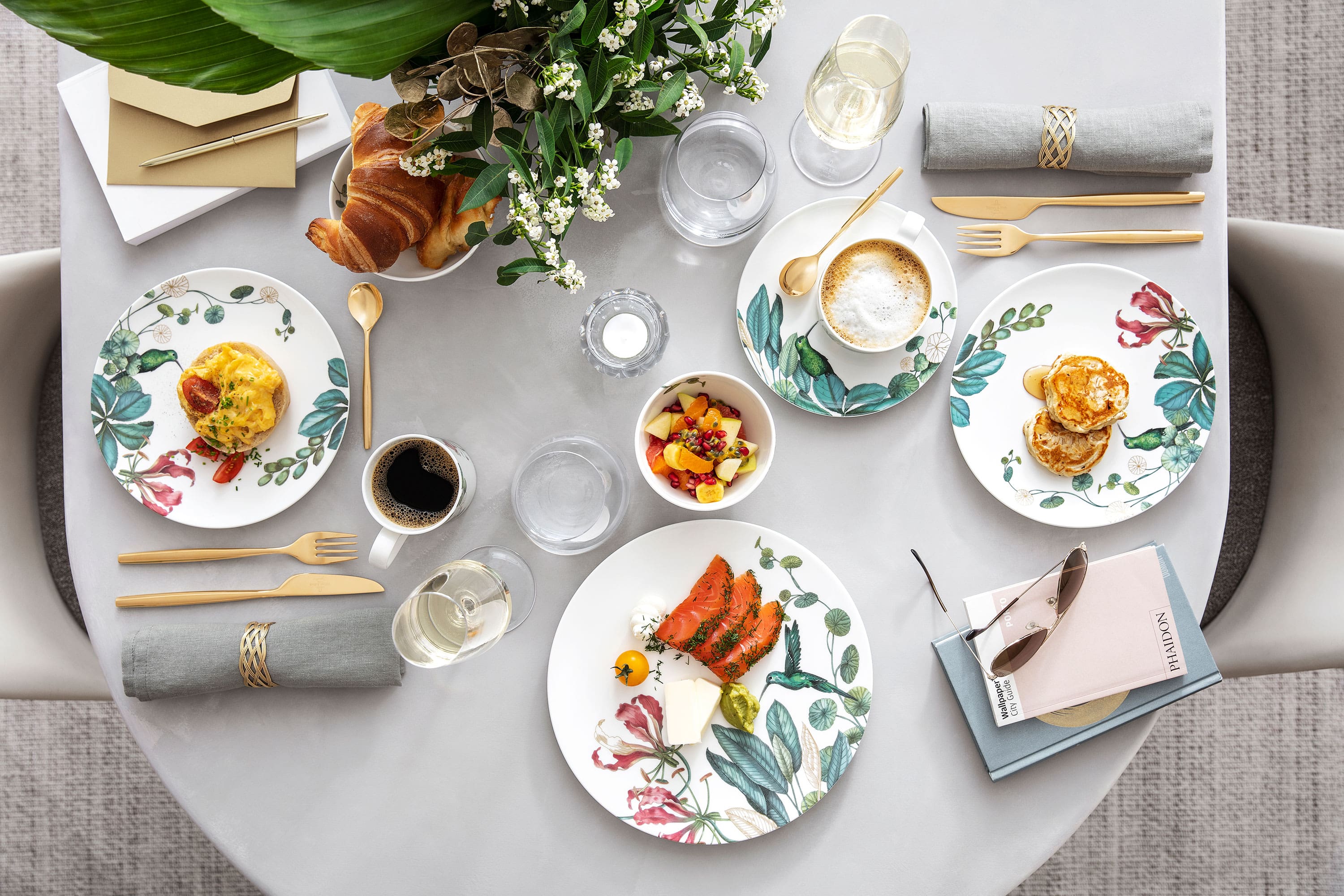 Table dressée pour un repas avec des assiettes fleuries, des aliments, des boissons, des serviettes et un petit bouquet de fleurs pour décorer la table.