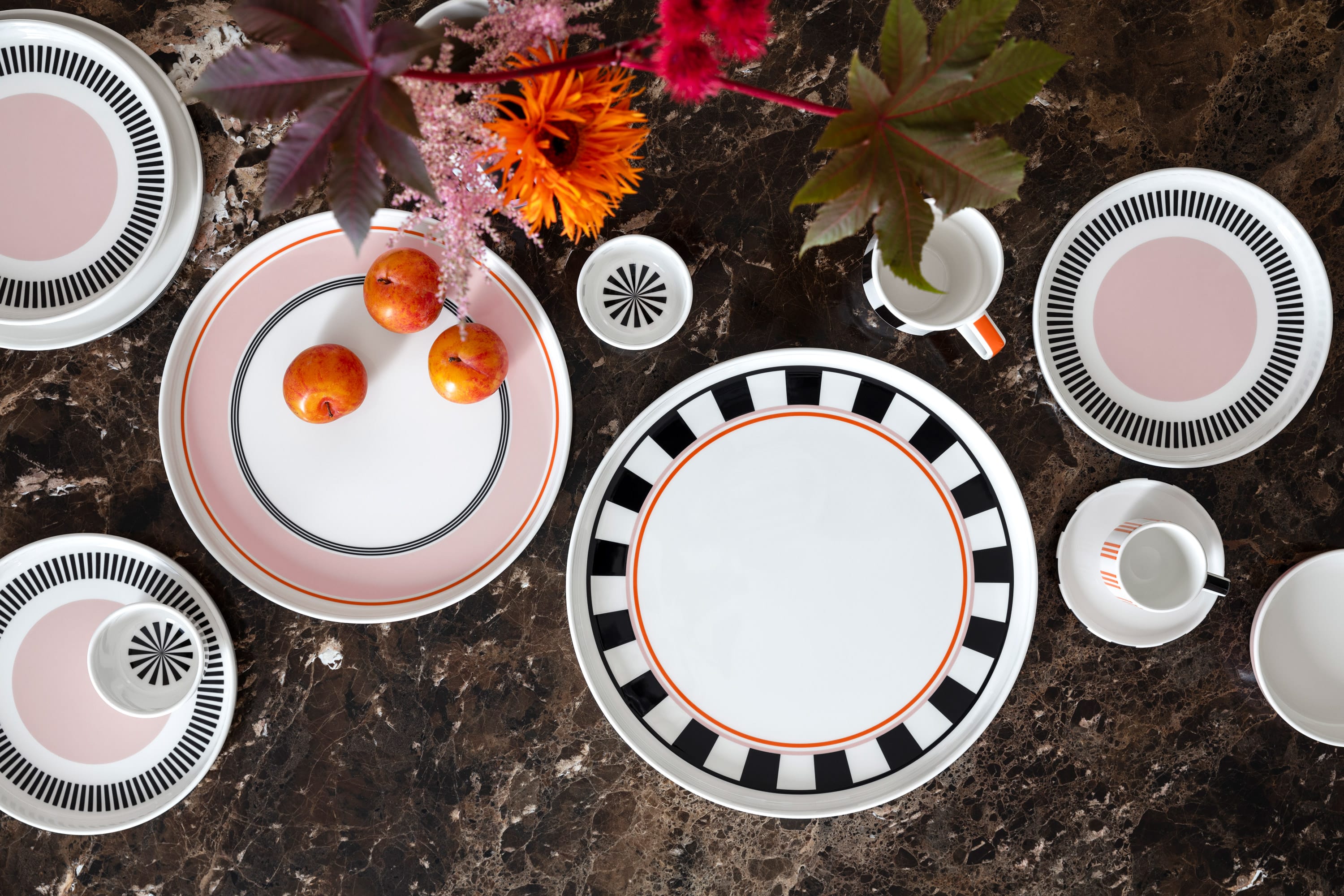 Service de table Memphis de Villeroy & Boch avec des assiettes décoratives, une tasse, des tasses et des prunes sur une surface en marbre brun décorée de fleurs brillantes.
