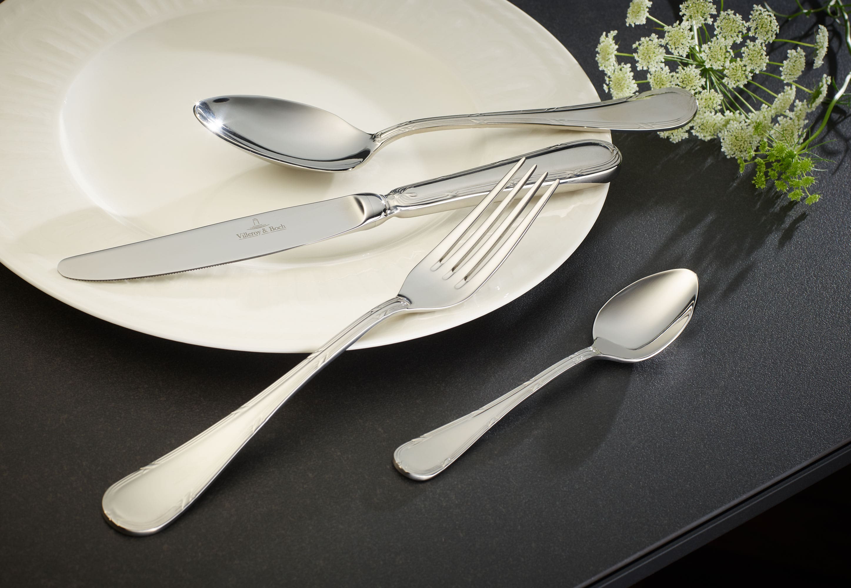 A white plate with flatware from the Kreuzband Septfontaine collection on a black surface.