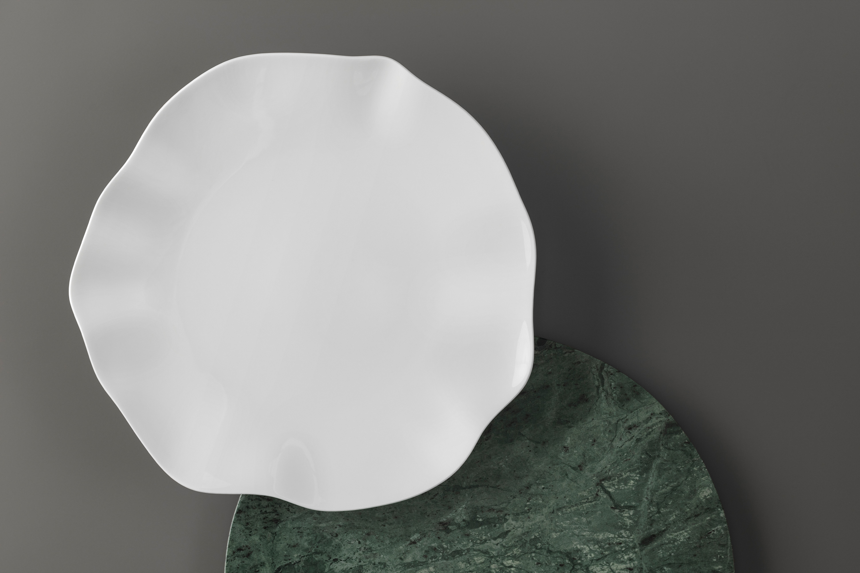 A white Pura plate with a scalloped edge on a dark green, round tray. A white Pura plate with a scalloped edge is placed on a dark green, round tray on a grey surface.