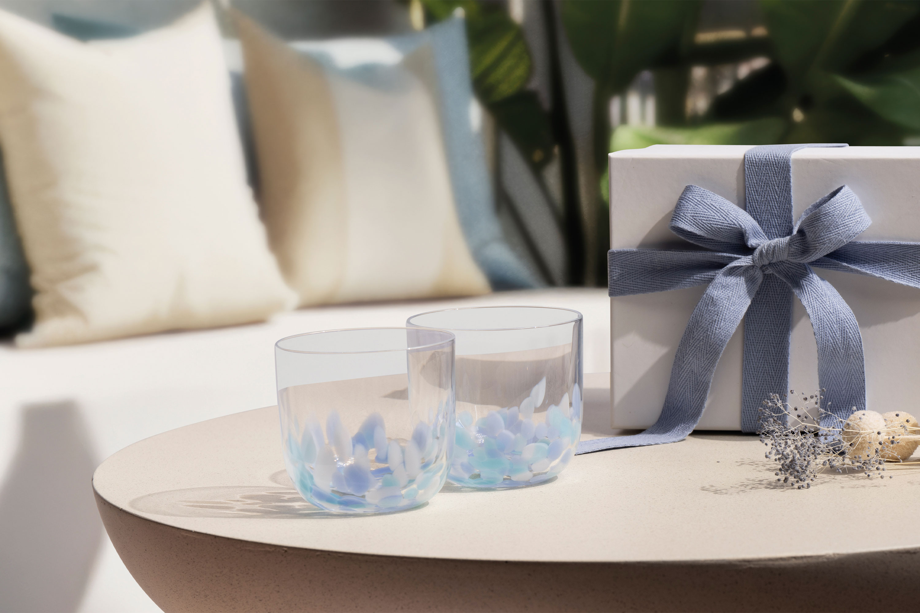 Two clear Like Aqua glasses and a white gift box with a blue ribbon on a round table; sofa and cushions in the background.