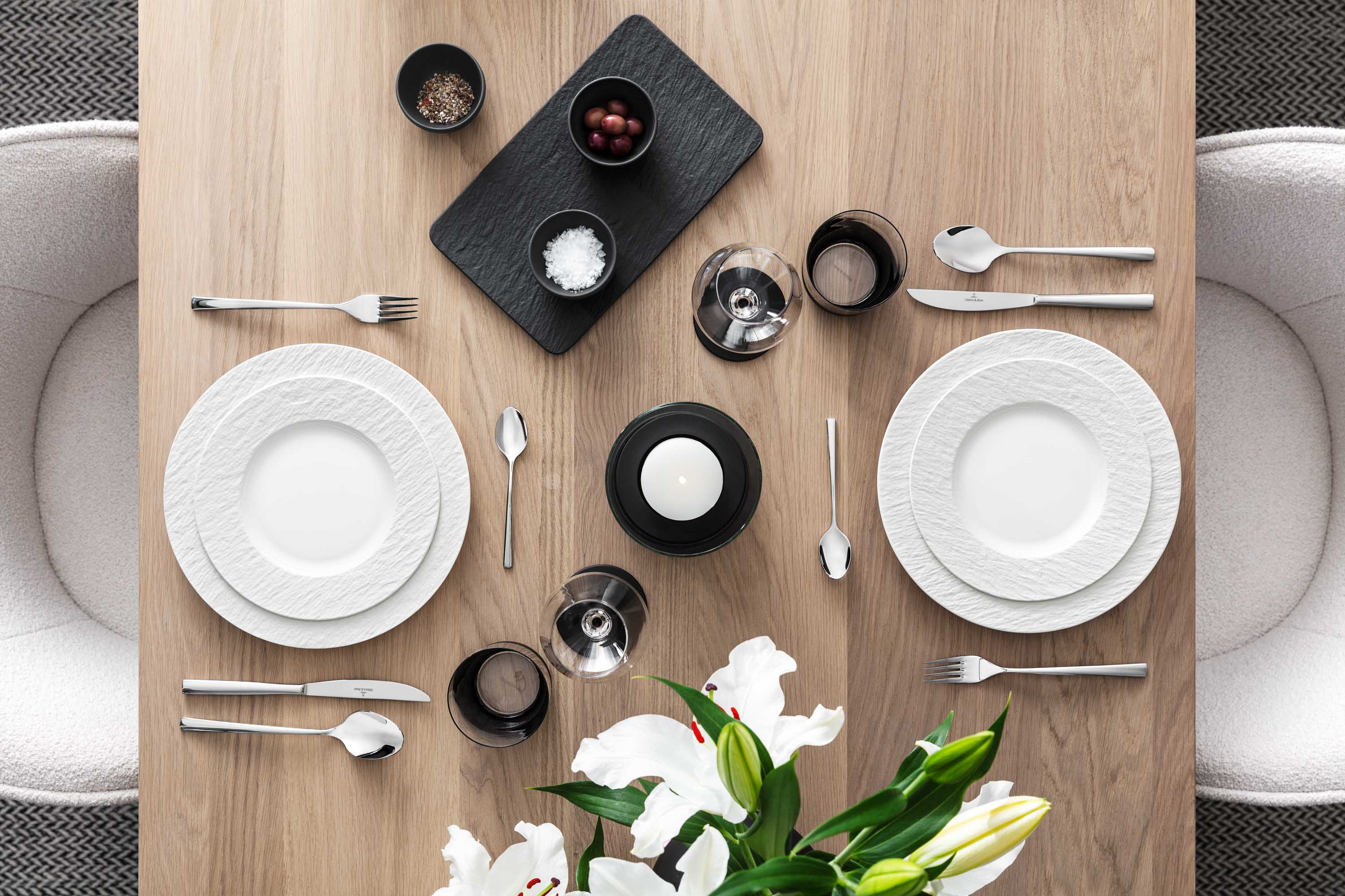 Elegant dining table with white Manufacture plates, Piemont cutlery and decorative flowers on a wooden surface.