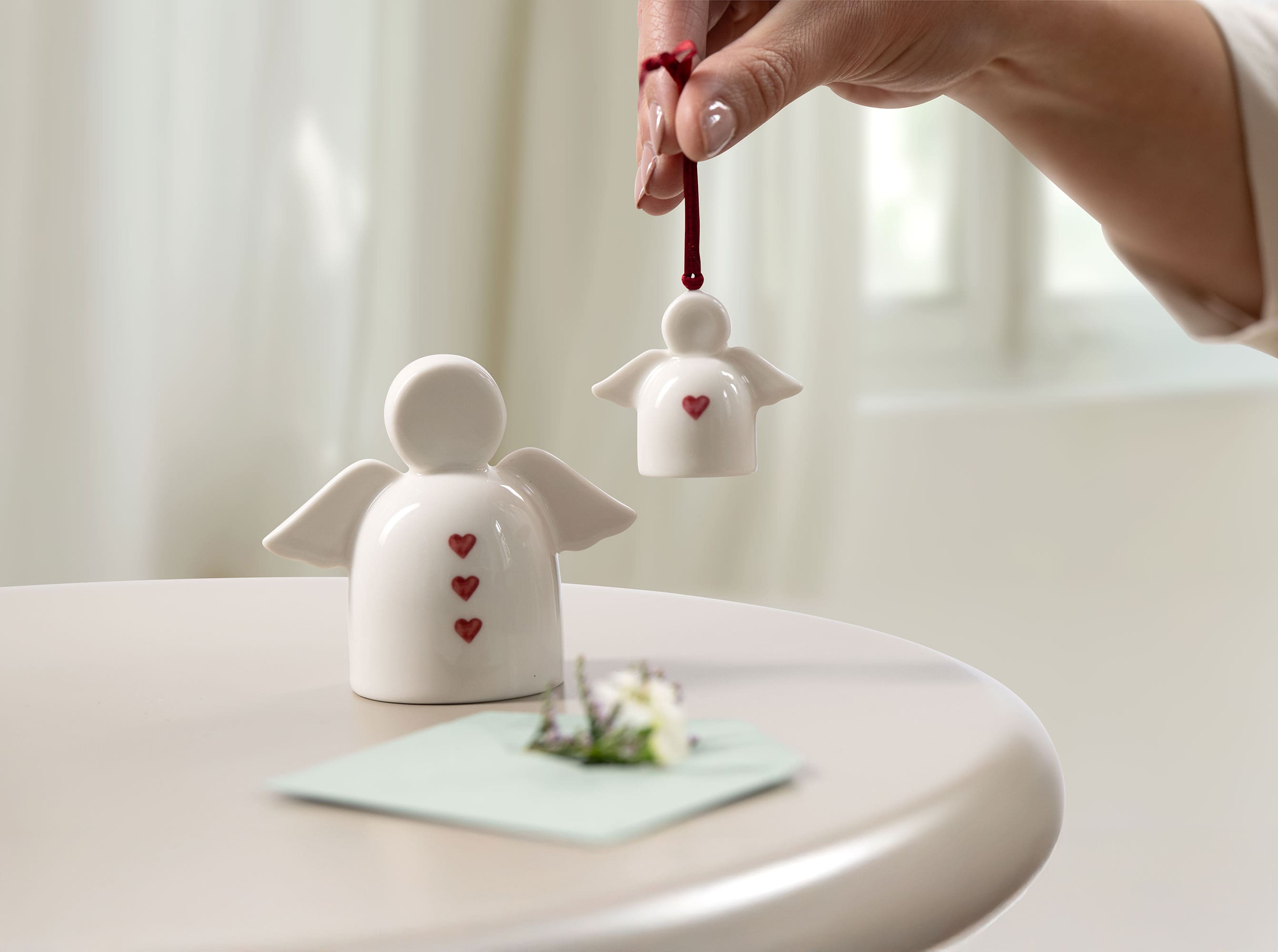 Adornos de ángeles con motivos de corazones rojos de with love como ideas para regalos Una mano sostiene un pequeño adorno de ángel de cerámica con motivos de corazones rojos de with love junto a una figura de ángel más grande de with love y una tarjeta sobre una mesa, que sirve como inspiración para regalos.