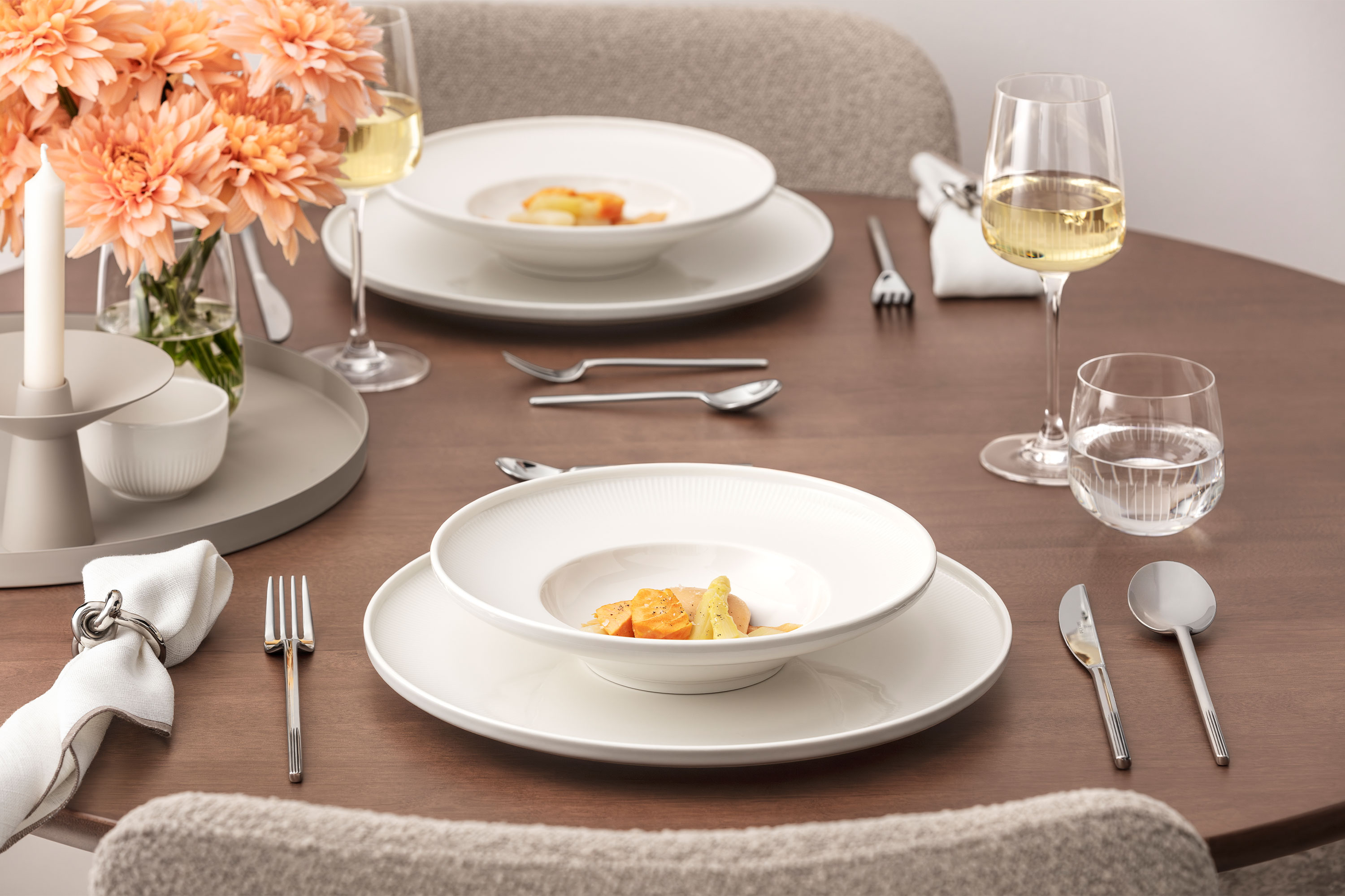 Una mesa bien puesta de la colección Afina con dos platos con platos gourmet, copas de vino, vasos de agua, cubiertos de plata, servilletas de tela y un jarrón con flores de melocotonero sobre una superficie de madera.