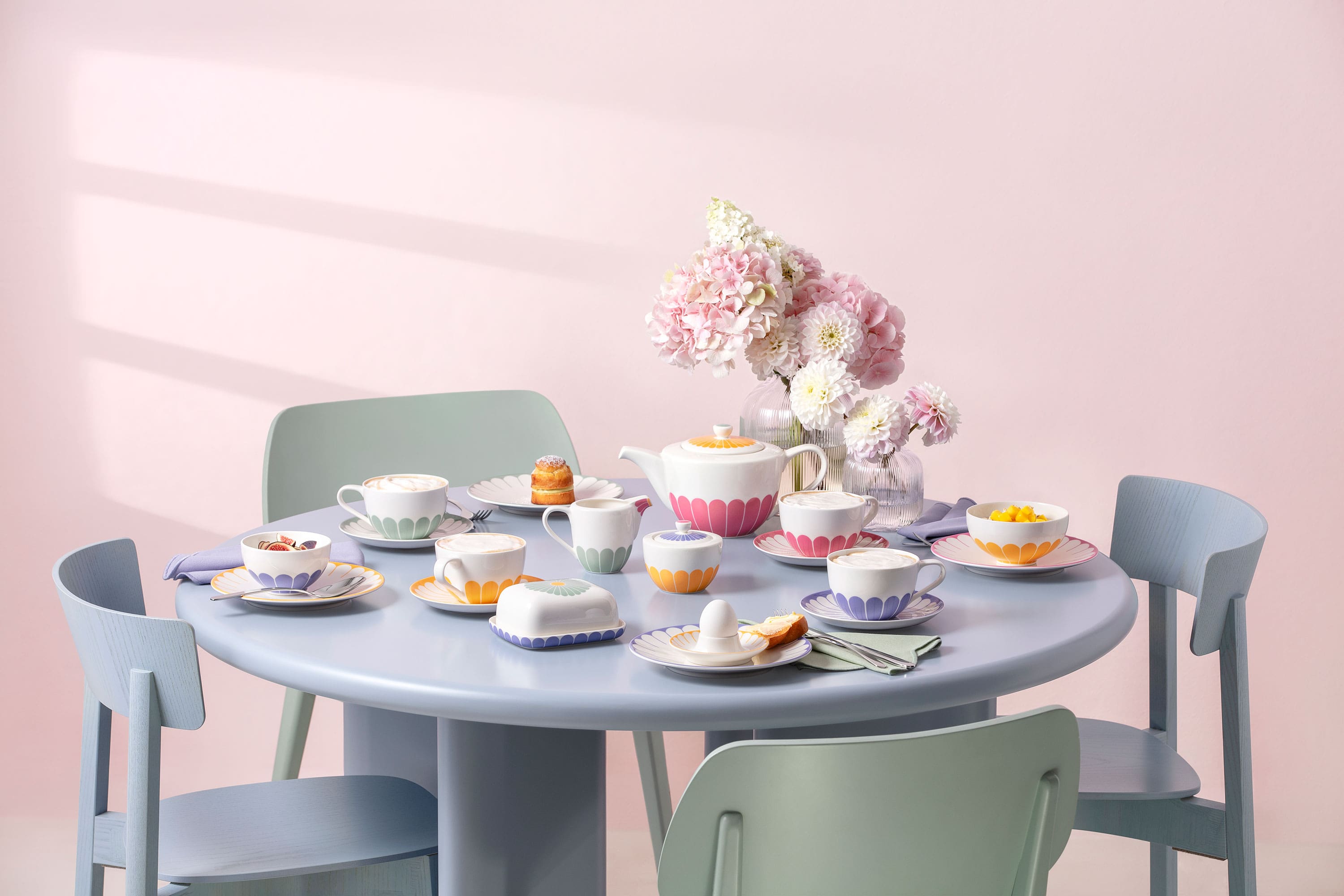 Floral table setting with tableware from Fleur for teatime Round pastel blue table with pastel chairs, set for tea, with cups and plates from Fleur with floral patterns, fine pastries, and a vase of pink flowers as the centerpiece.