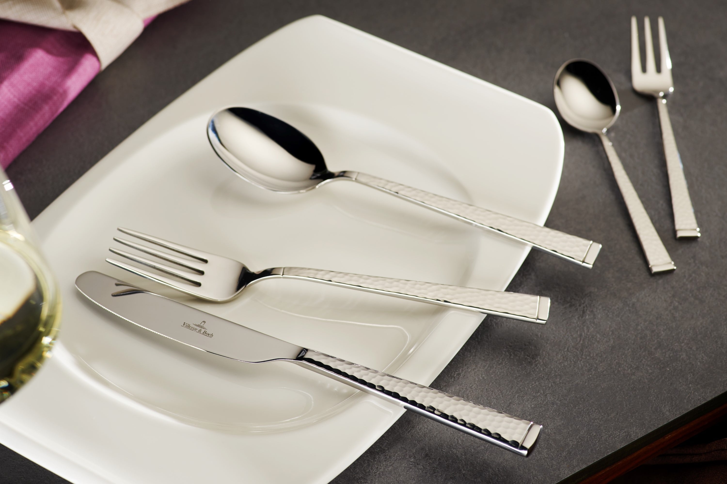 A white rectangular plate from Villeroy & Boch Blacksmith features a shiny fork, knife, and spoon on a gray surface. Next to it are a smaller spoon and a fork from the Blacksmith collection. A wine glass and a pink napkin are partially visible.