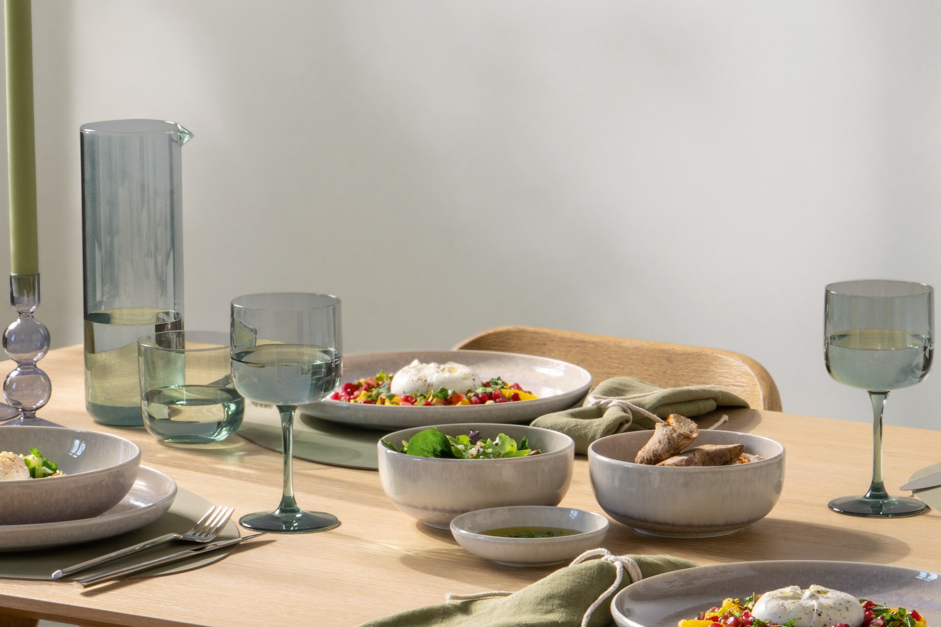 Like Carafe Table Set with Perlemor A modern dining table with a carafe, gray tableware, glasses, food, and green napkins in natural light.