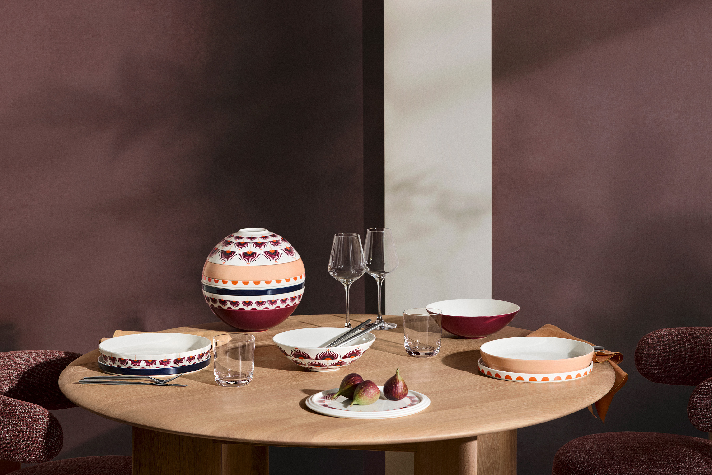 La Boule Kyoto tablesetting A round wooden table set with patterned ceramic tableware from La Boule Kyoto, two wine glasses, and a decorative vase against a brown background.