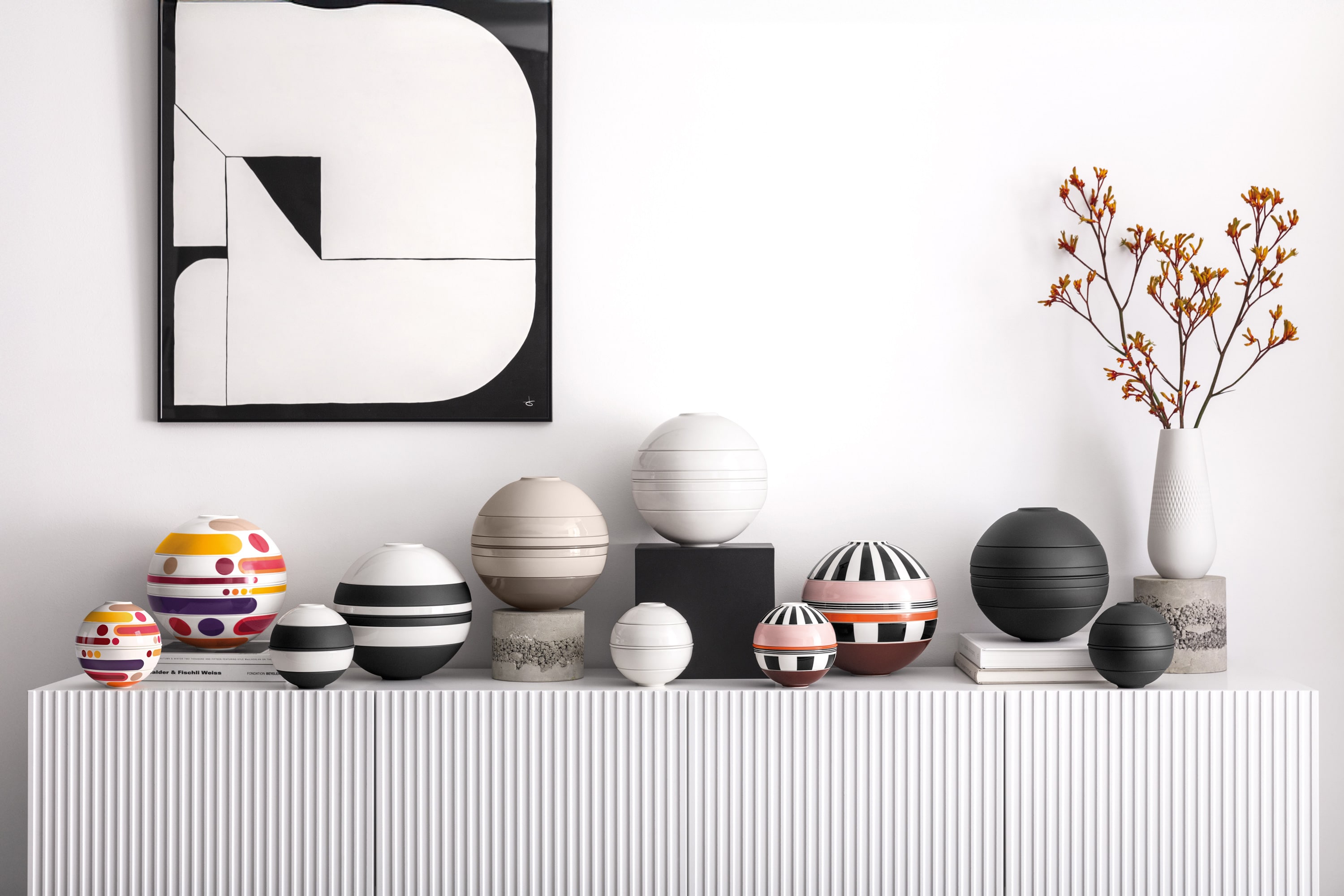 Various La Boule pieces in different sizes and colors A collection of colorful, striped, and patterned spherical La Boule pieces stands on a white console beneath abstract art and a vase of orange flowers.