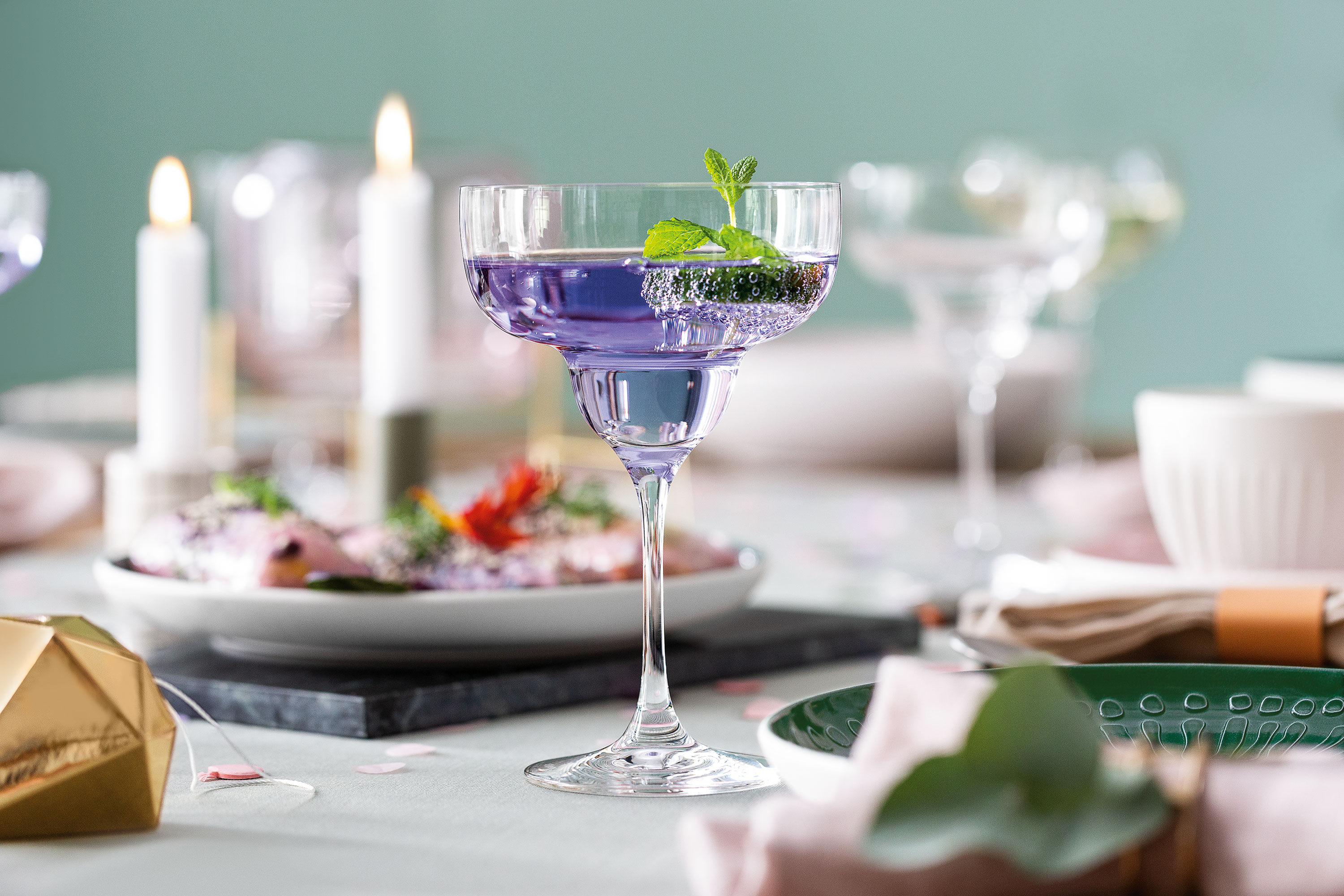 A purple cocktail, garnished with mint, in a Purismo Margarita glass on a set table, surrounded by plates, candles, and decorative objects.