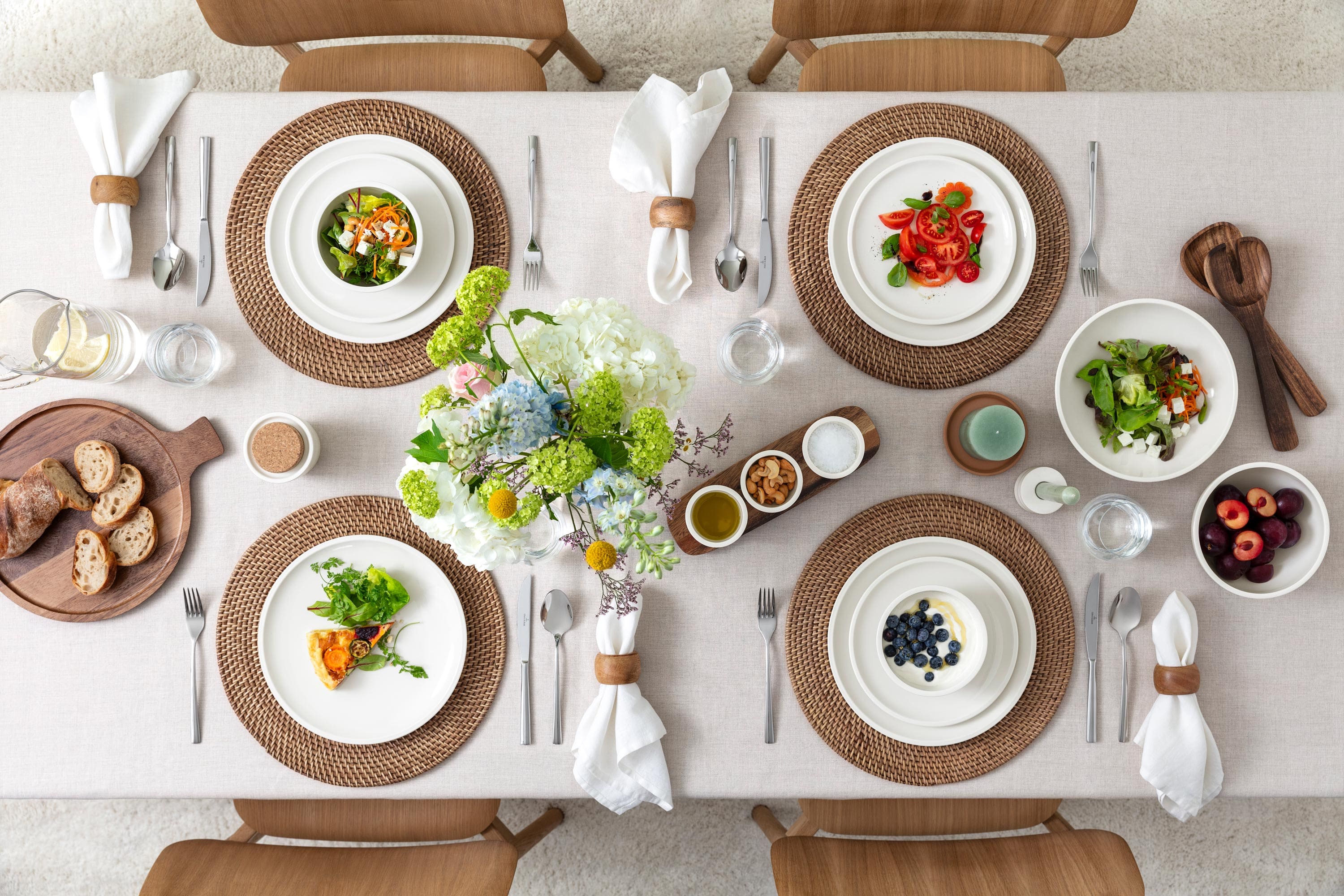 Vue de dessus d'une table à manger joliment dressée avec des salades, des fruits, du pain, des verres d'eau et des fleurs au centre sur une nappe beige.
