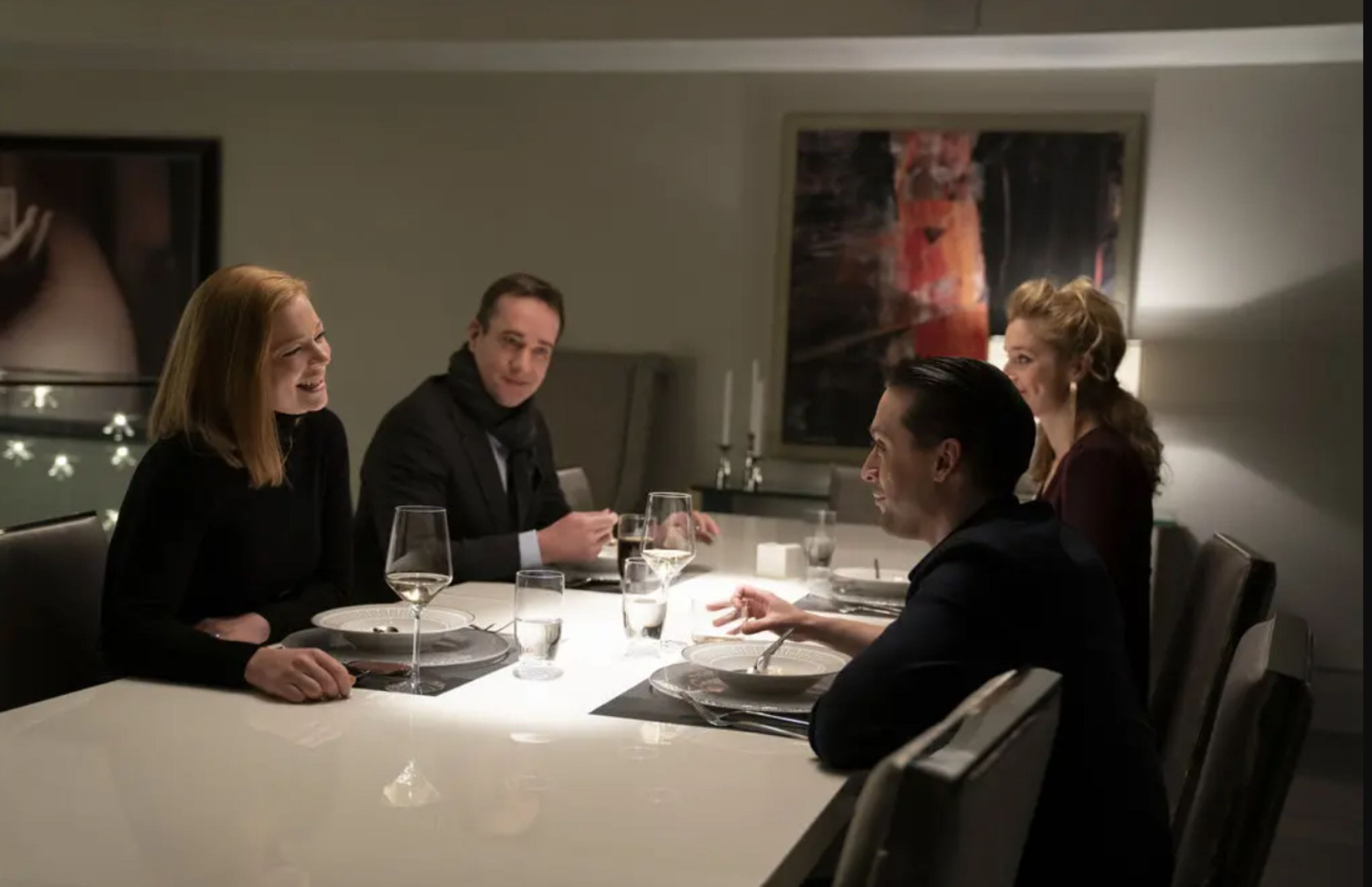 succession-as-seen-on A group of four people sit around a modern dining table with white plates and glasses, warmly engaged in conversation. The room is dimly lit and stylish, with abstract art on the wall, conveying a sophisticated, intimate gathering.