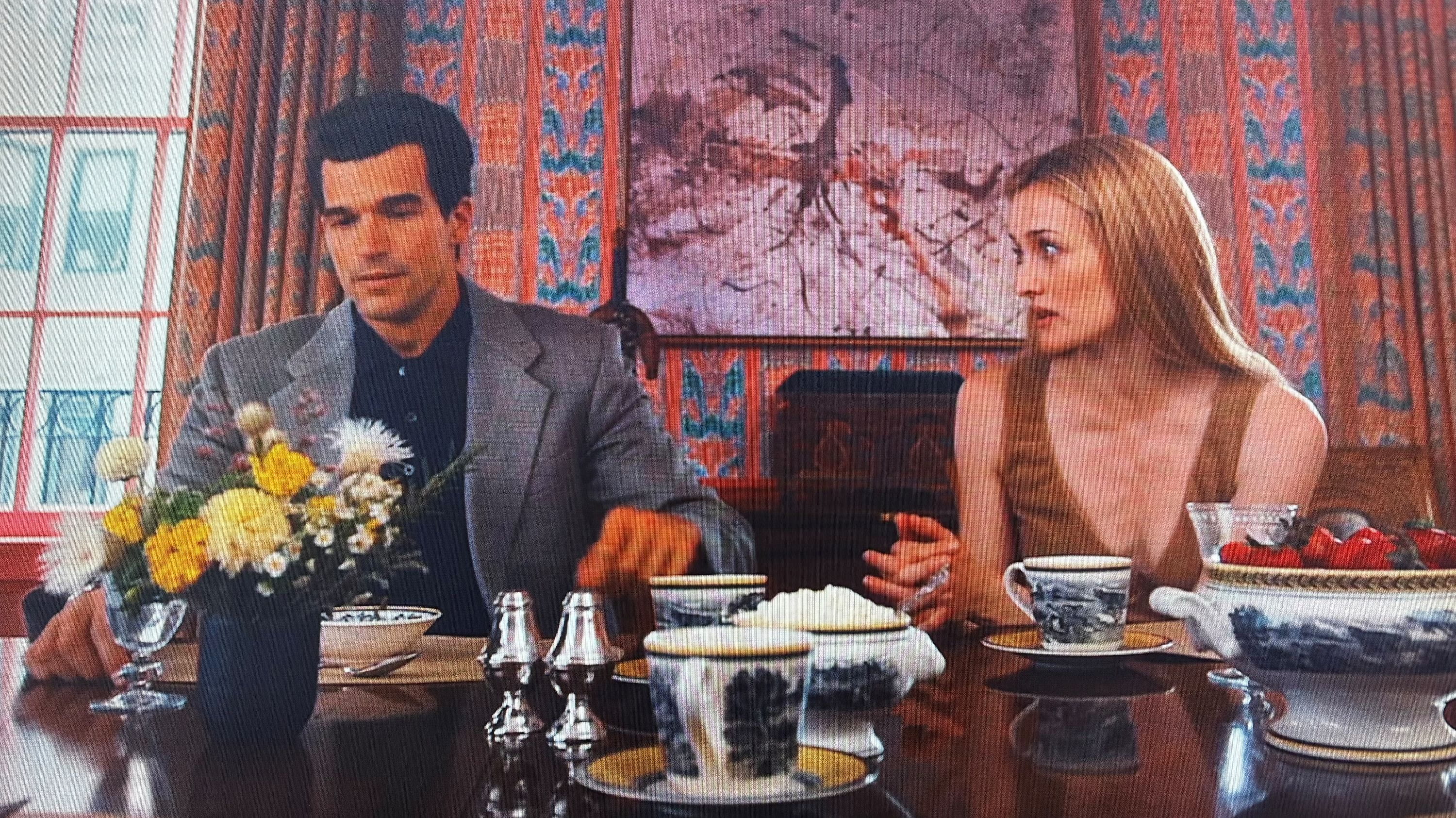 A man and woman seated at a dining table in an elegantly decorated room, engaging in conversation. The table is set with flowers, china, and silverware.