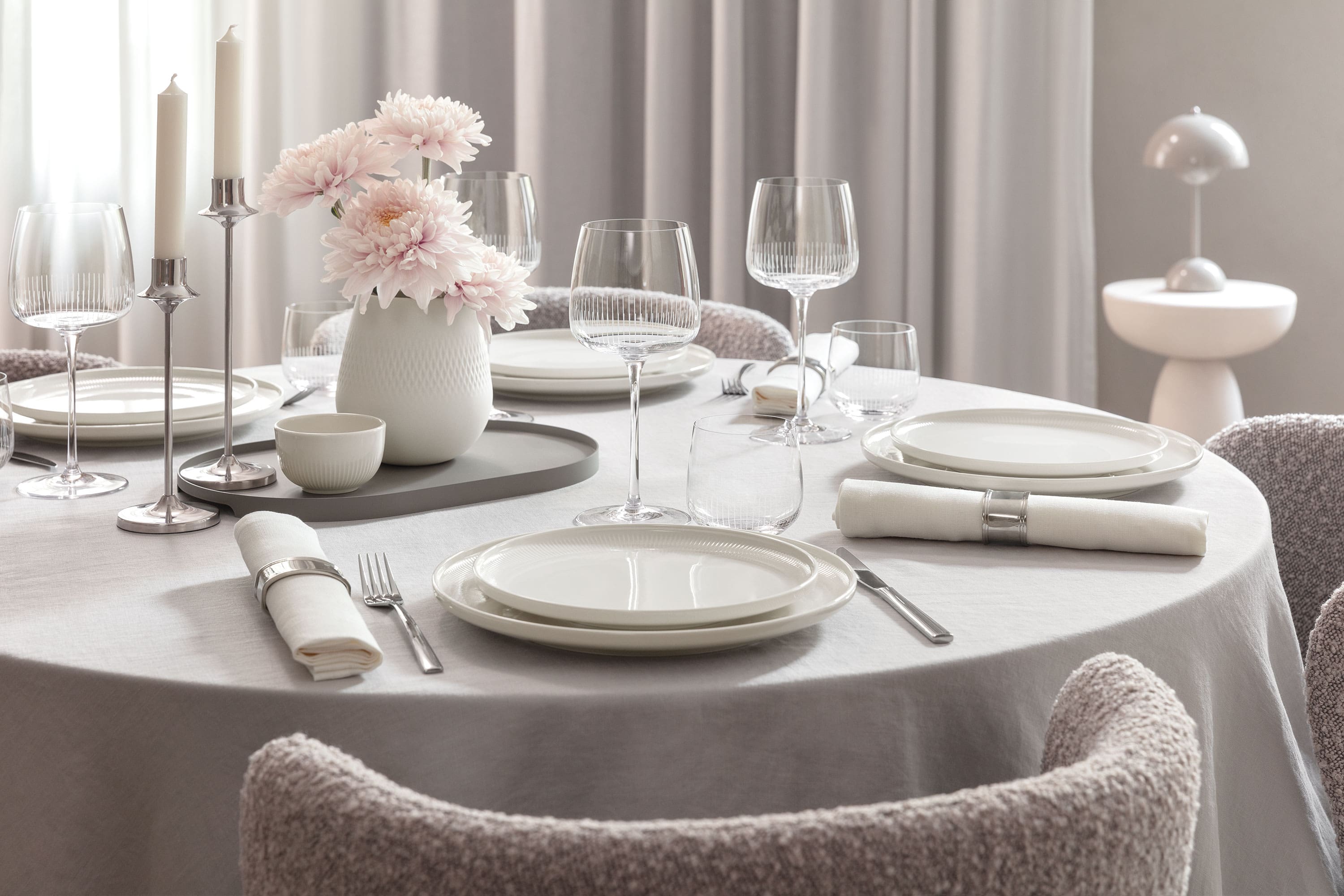 Runder Esstisch mit weißen Afina Tellern, Afina Gläsern, Besteck, Servietten und einer Vase mit rosa Blumen.