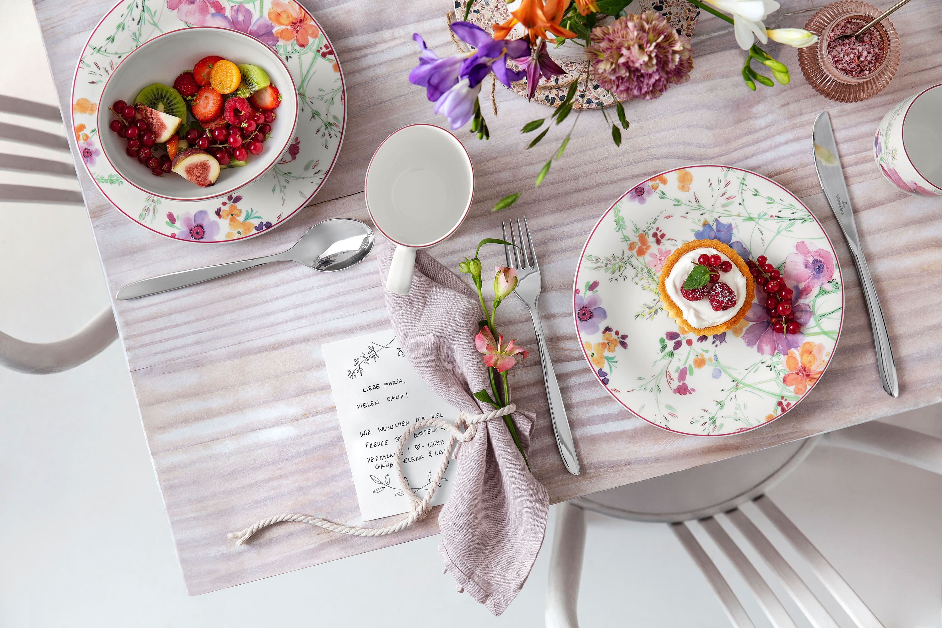 A table laid with a floral pattern with plates, cutlery, a napkin, a small dessert and a bowl of fruit on a light-coloured tablecloth.