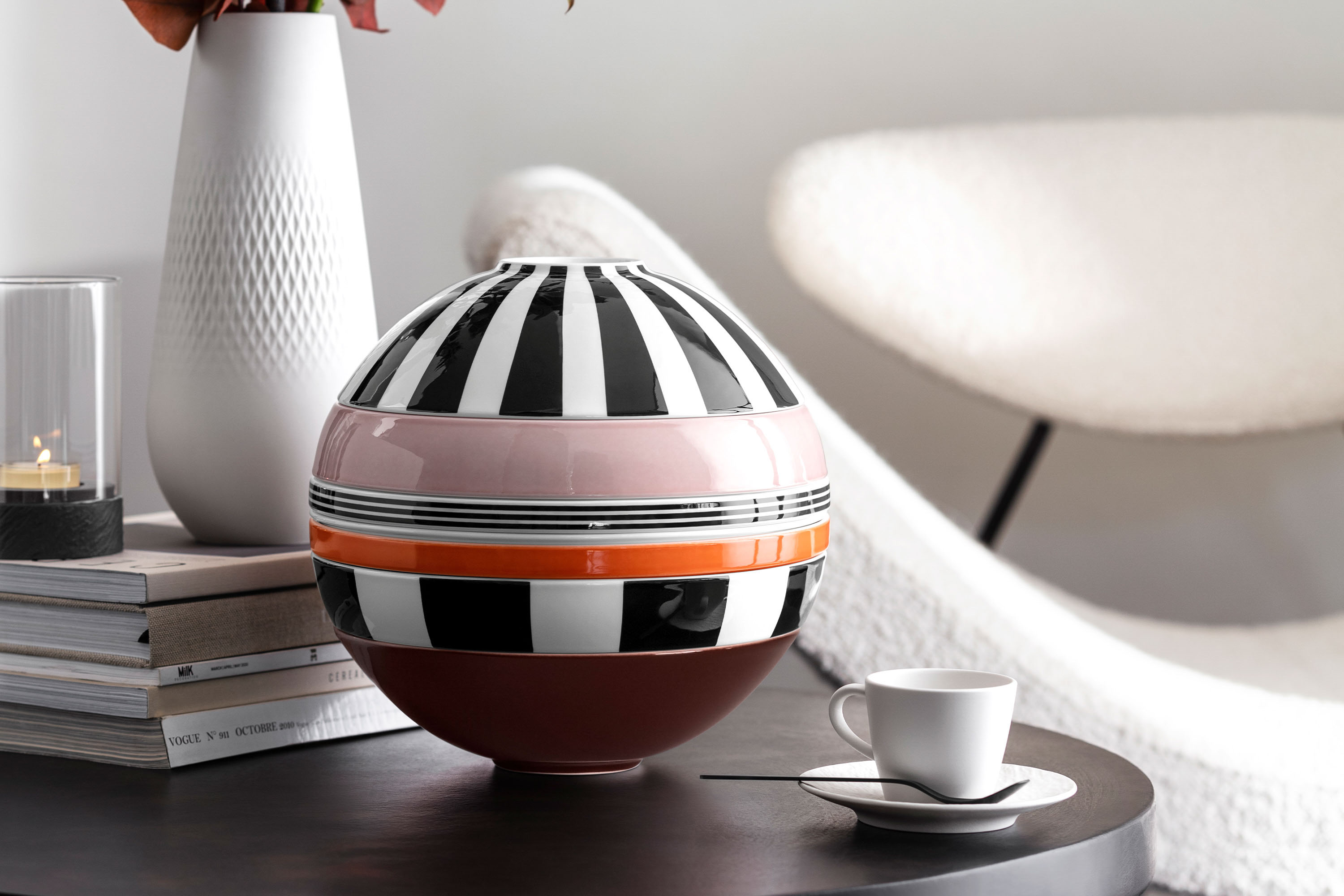 La Boule Memphis on a side table Memphis La Boule with monochrome stripes and a horizontal stripe in pink and orange on a table next to a white cup and saucer, stacked books, and a textured white vase. There is a modern chair in the background.