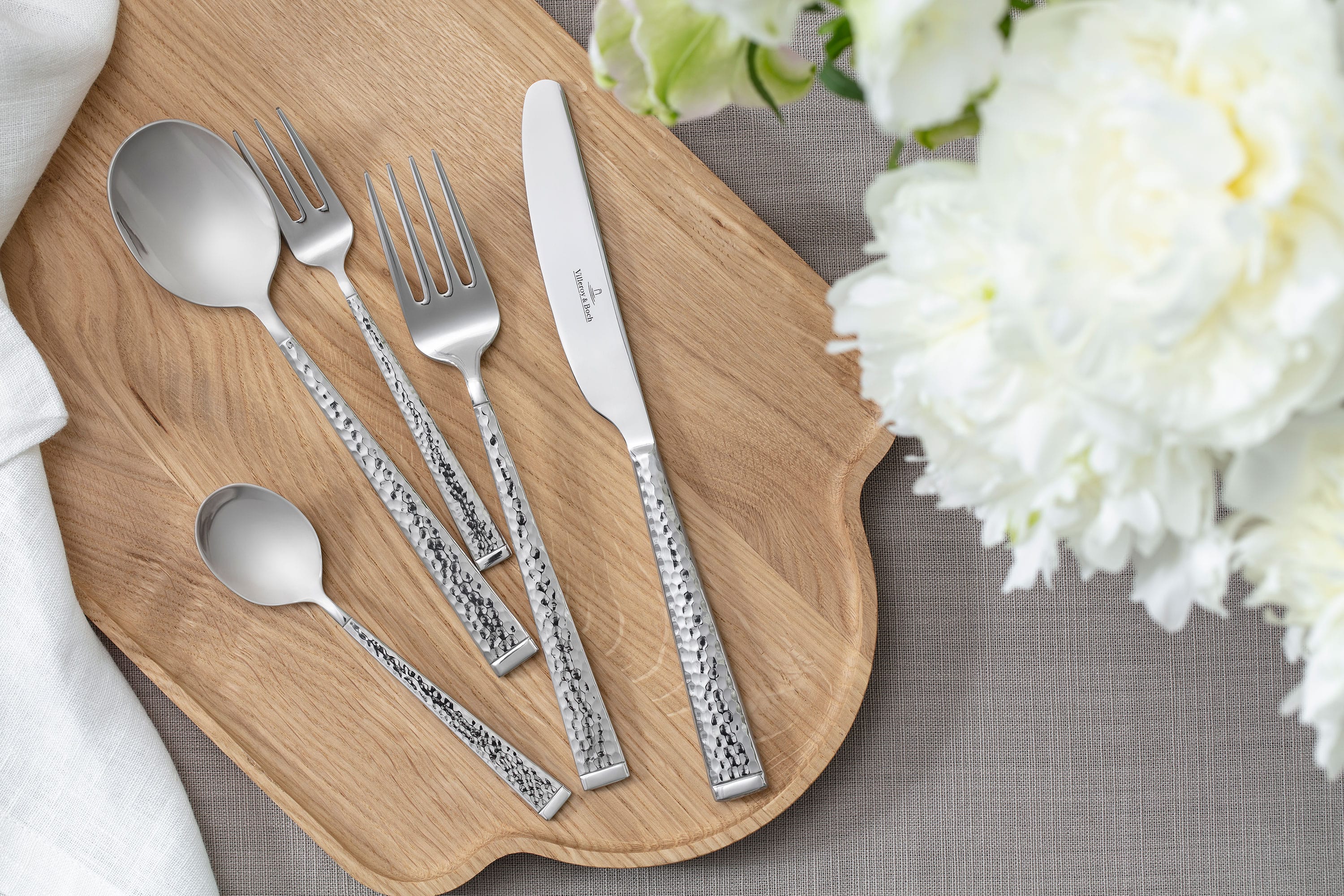 Blacksmith Cutlery – Arranged on a wooden board Top-down view of Blacksmith cutlery – tablespoon, table fork, table knife, coffee spoon, and dessert fork – neatly arranged on a wooden board. Surrounded by delicate white floral decor. A clean, natural, and elegant aesthetic.