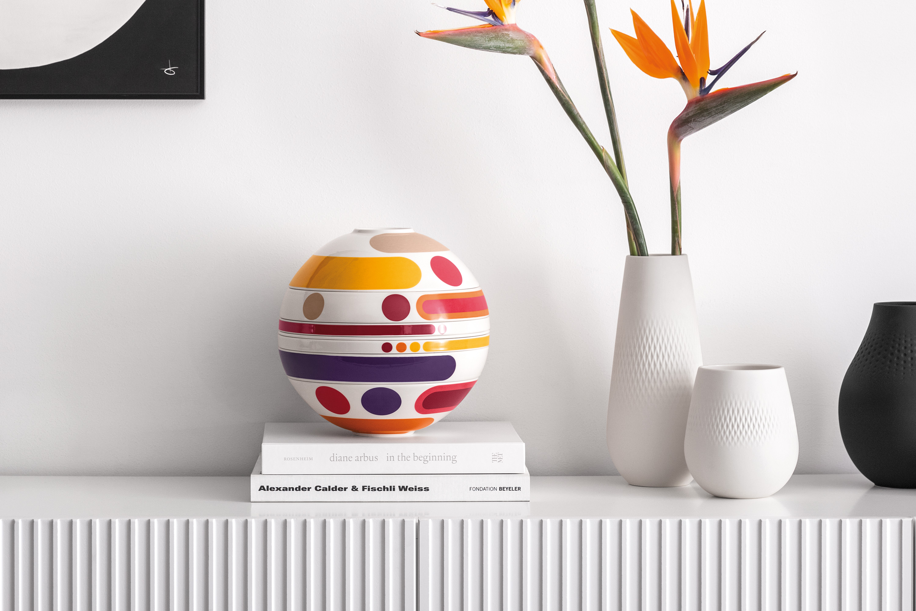 A colorful La Boule Miami on books next to white vases and bird of paradise flowers on a white shelf.